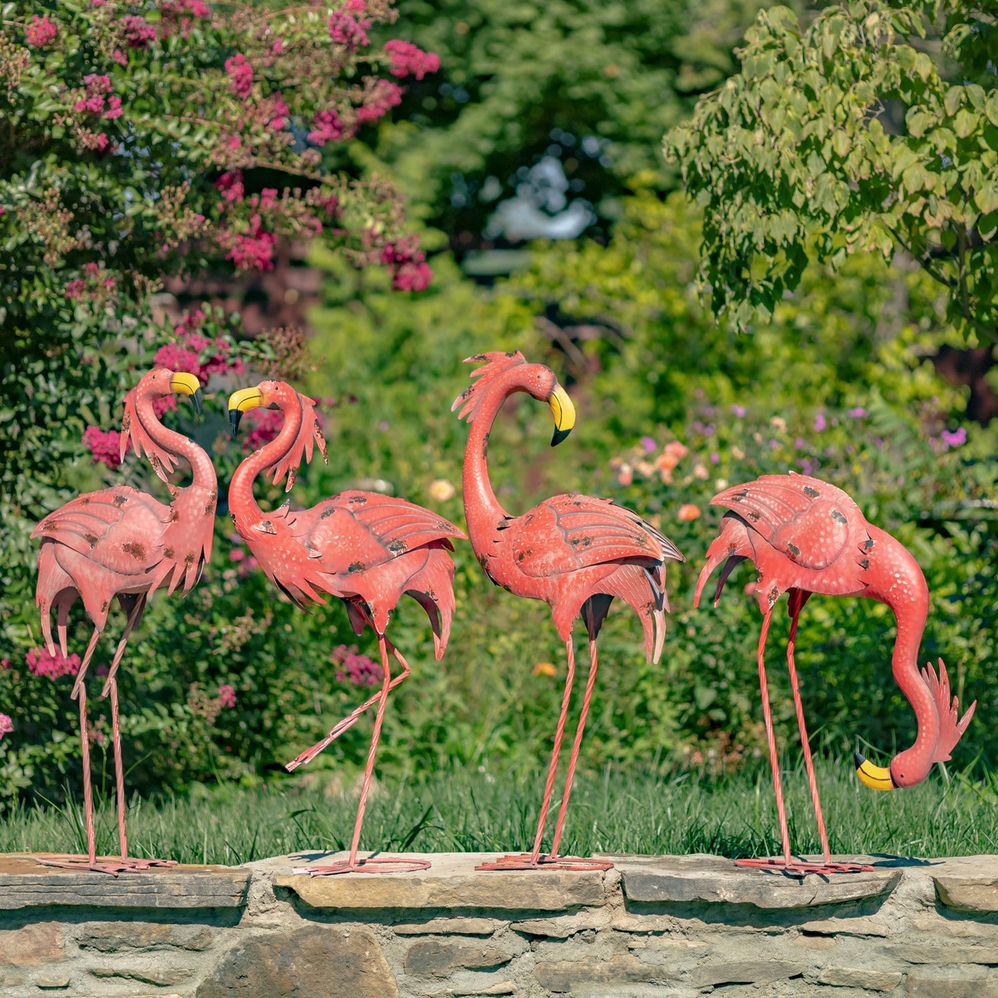 Four pink flamingo garden statues in different poses, displayed on a stone wall in a lush green garden with pink flowers.