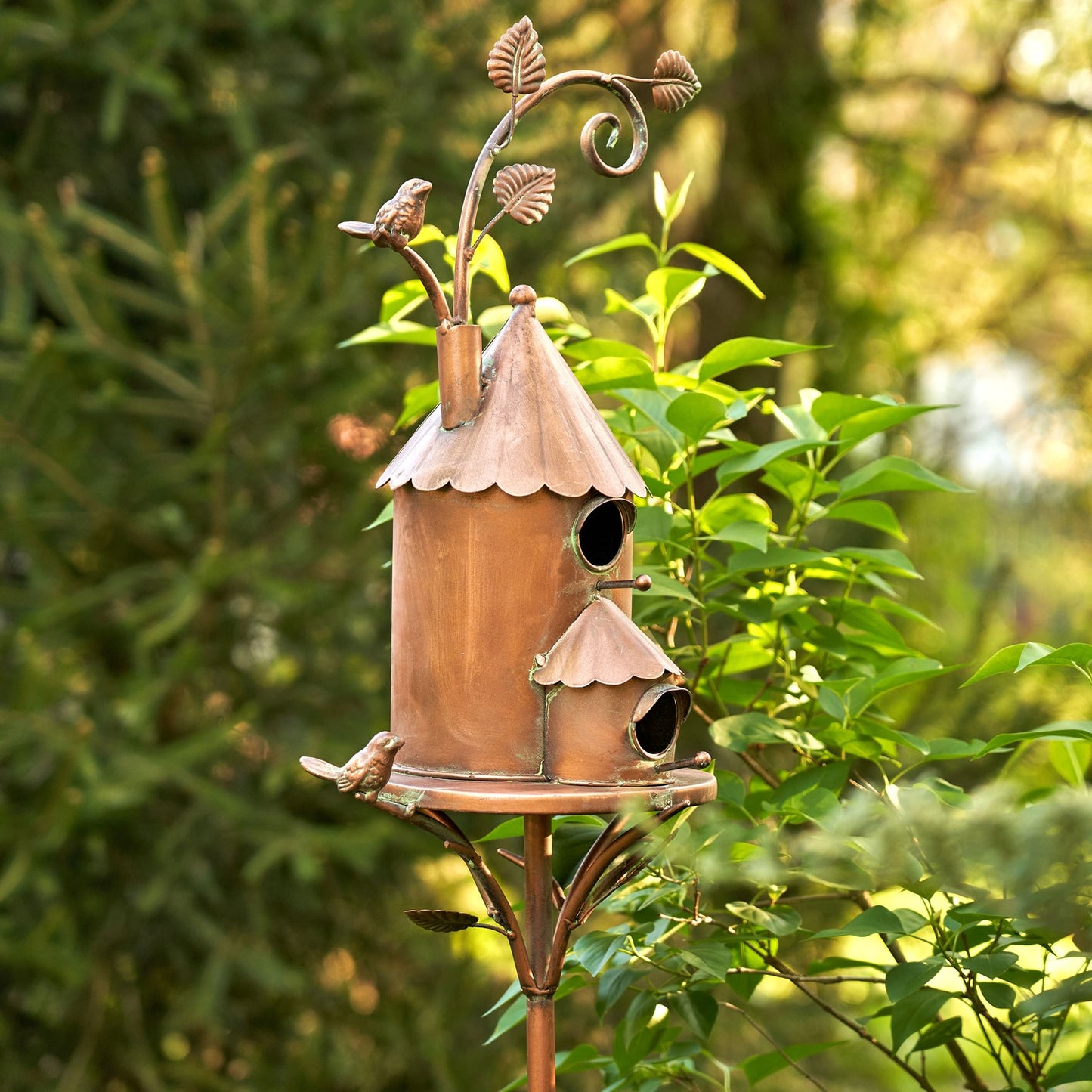 A decorative copper birdhouse with two tiers, perched on a metal stand with bird-shaped perches and a leaf scroll finial, photographed outdoors among green garden plants.