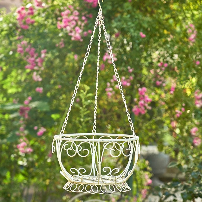 Set of two antique white iron hanging basket planters suspended by decorative chains and filled with blooming flowers on a cottage-style porch.