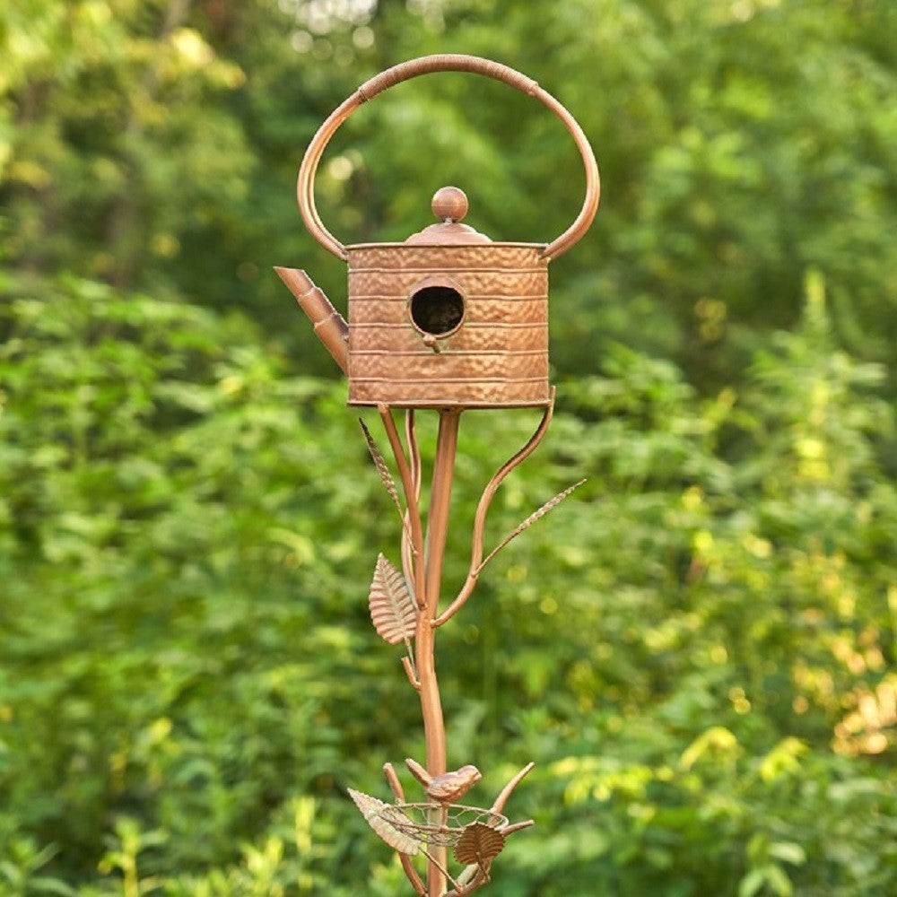A decorative metal garden stake shaped like a birdhouse watering can, with a copper-toned birdhouse top, loop handle, spout, and leafy stem details, photographed outdoors against a blurred green foliage background.