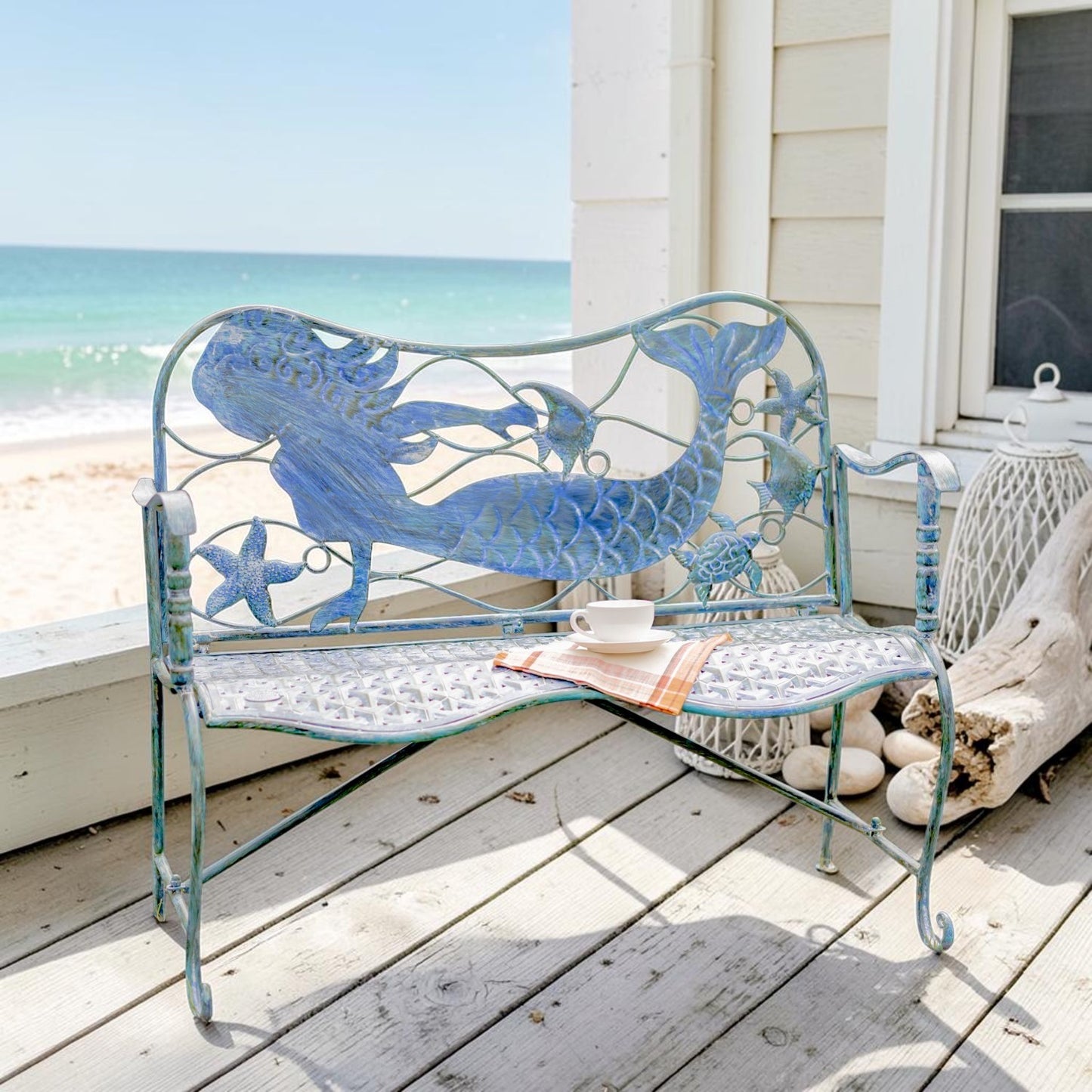 A decorative metal beach bench painted in light blue and turquoise, featuring a mermaid and starfish design, set on a white wooden deck with the ocean and a house exterior in the background.