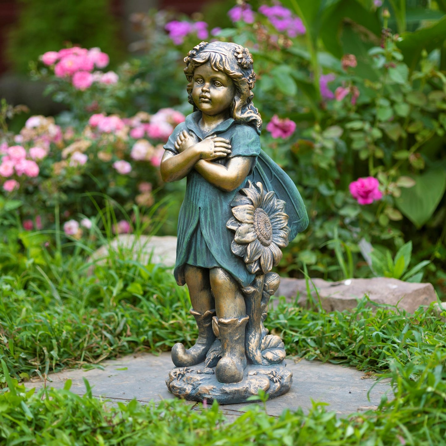 A bronze-finished garden statue of a young girl with arms crossed, holding a large sunflower, displayed on a stone path in a lush garden with pink flowers in the background.