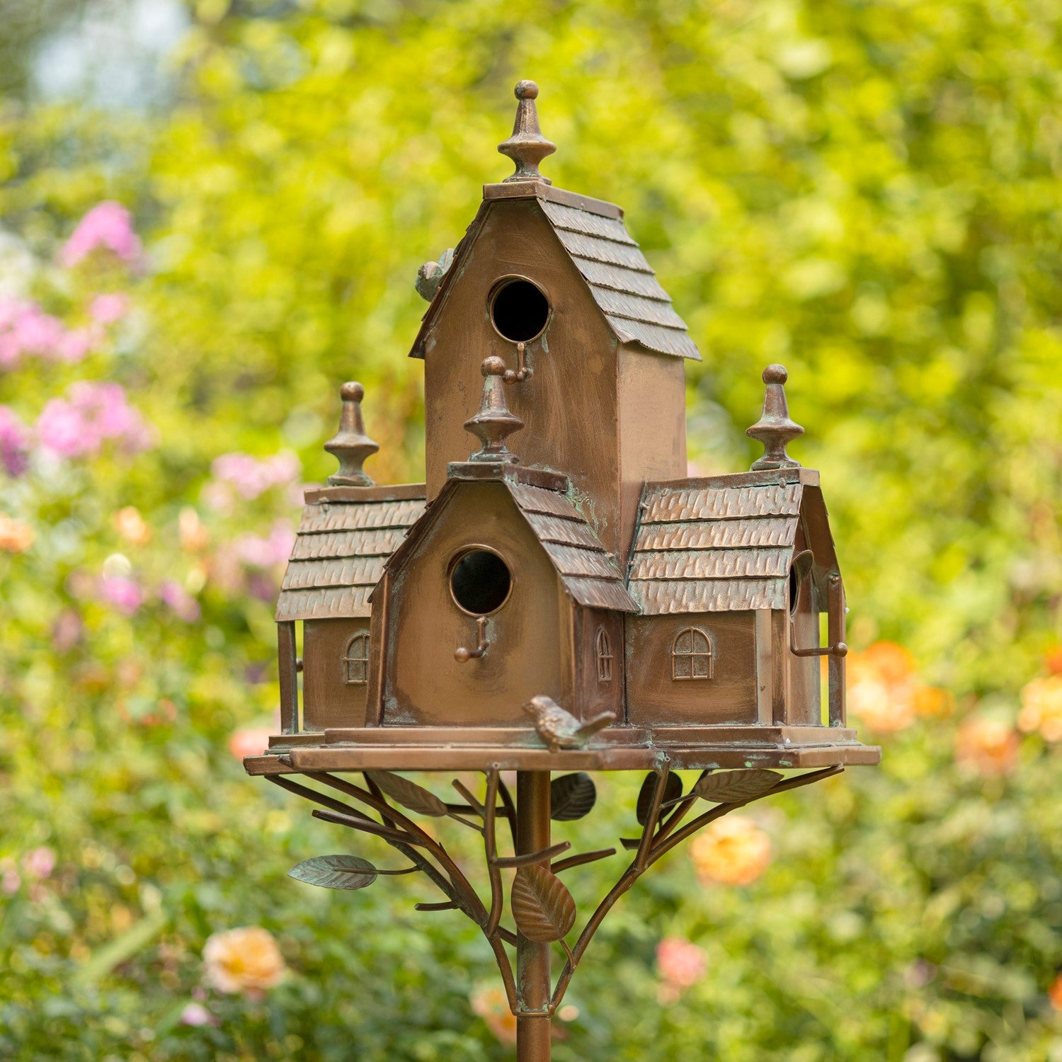 A multi-tiered birdhouse with a rustic brown and bronze finish, mounted on a decorative metal stand with leaf details, set against a blurred green garden background with pink flowers.