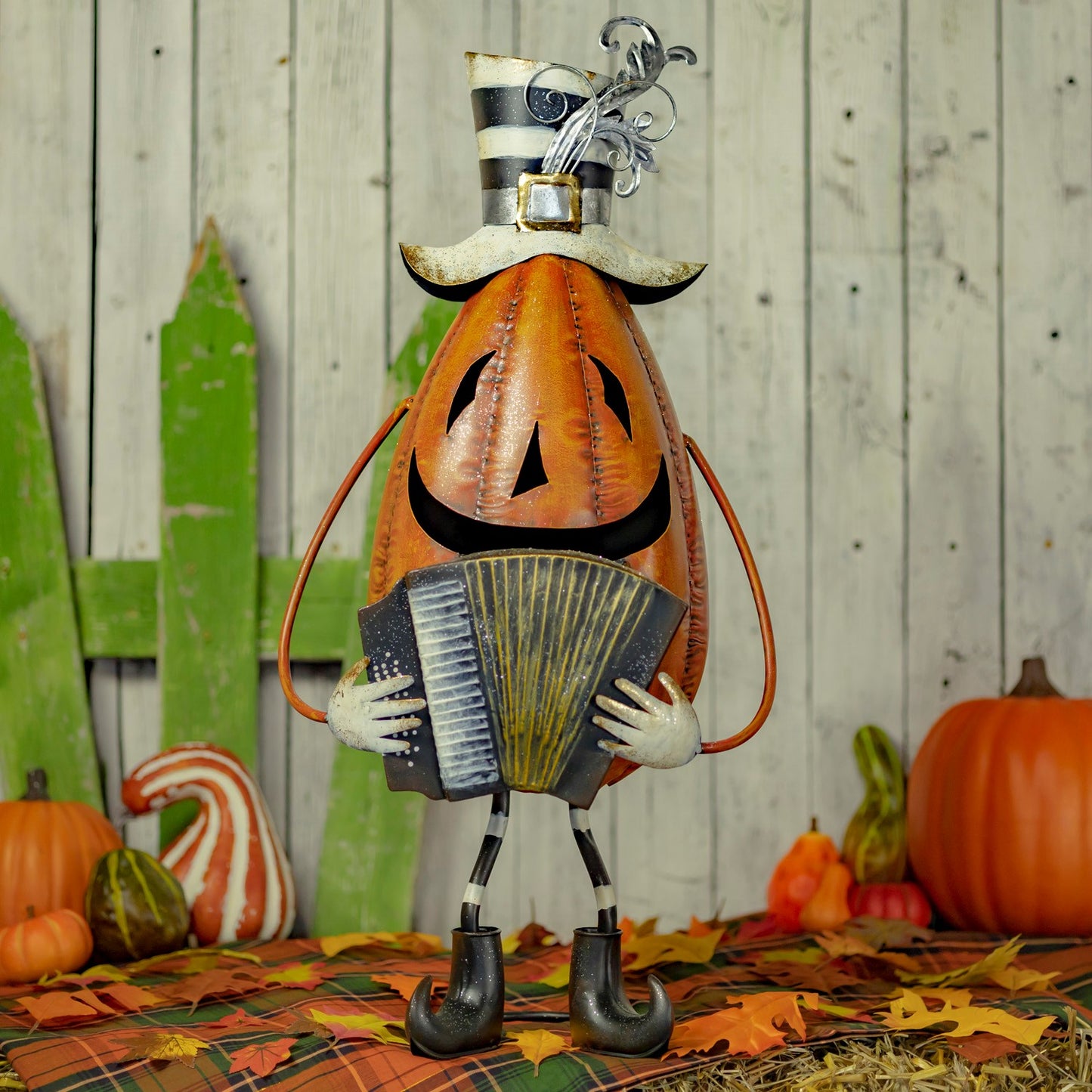 A Halloween decorative pumpkin figure wearing a black-and-white striped top hat and playing an accordion, posed on a plaid blanket with pumpkins, fall leaves, and a rustic wood plank backdrop.