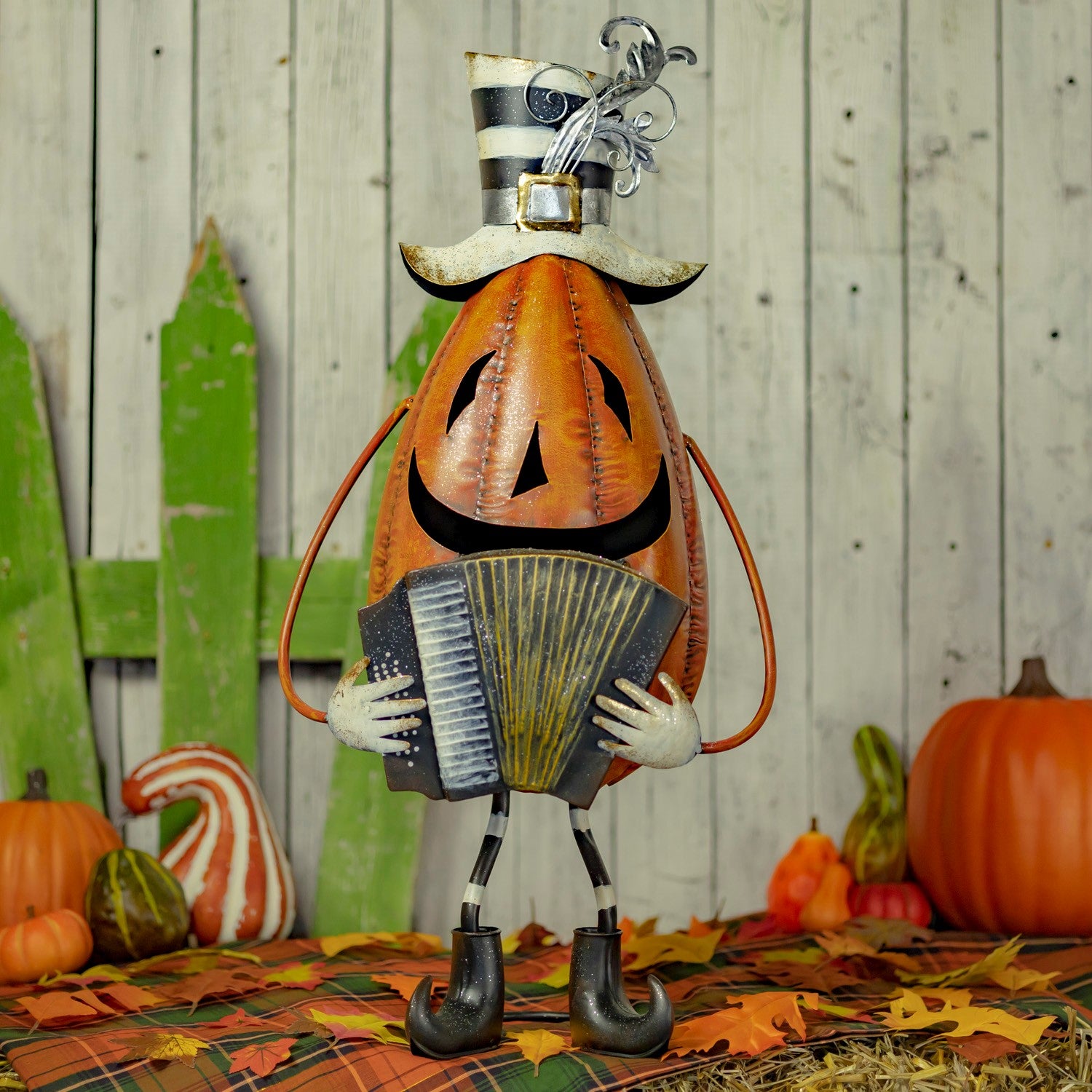 A Halloween decorative pumpkin figure wearing a black-and-white striped top hat and playing an accordion, posed on a plaid blanket with pumpkins, fall leaves, and a rustic wood plank backdrop.