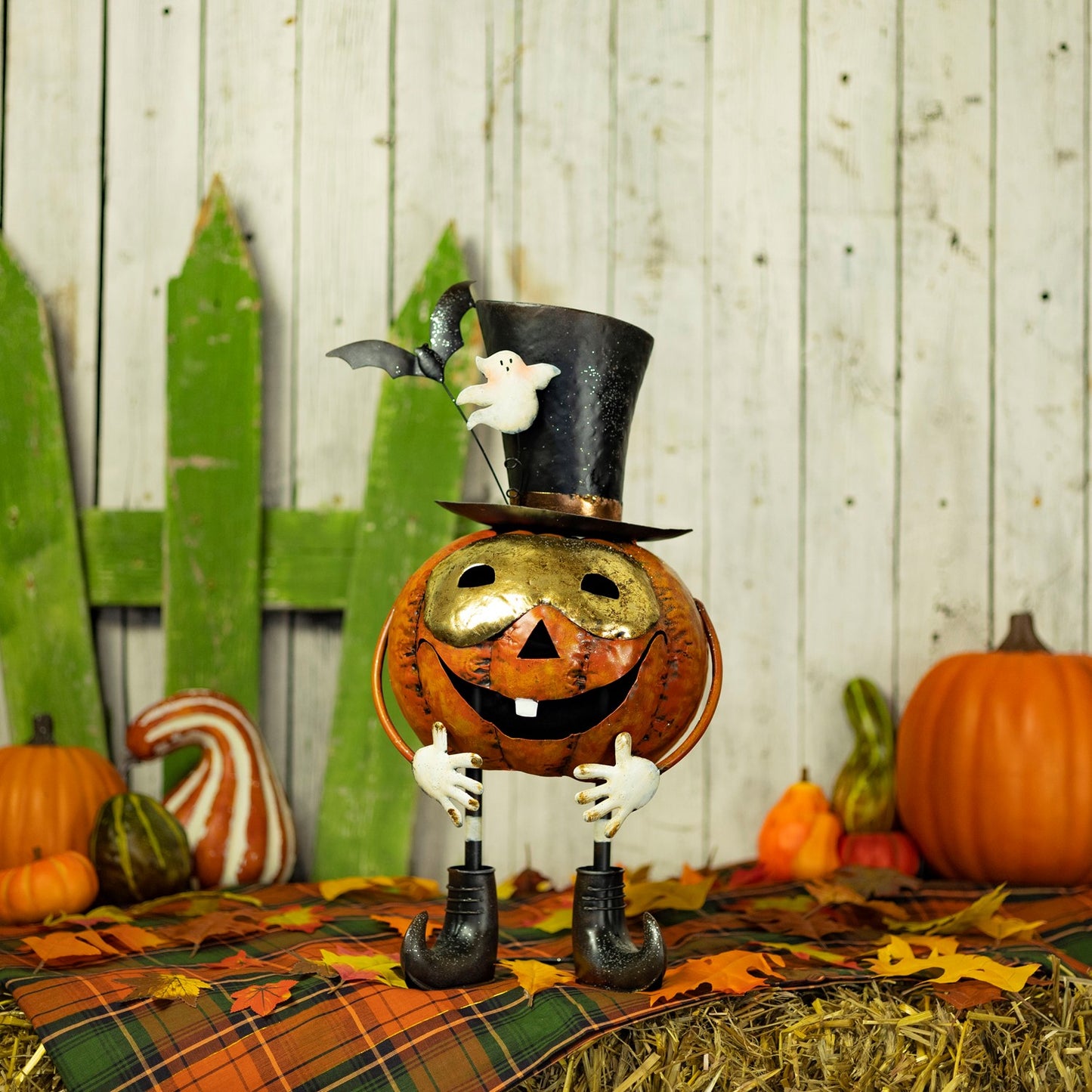 A Halloween decorative pumpkin figure with a gold face, black top hat, white ghost, and black umbrella-shaped legs, posed on a plaid blanket with hay, fall leaves, pumpkins, and a rustic wood plank backdrop.