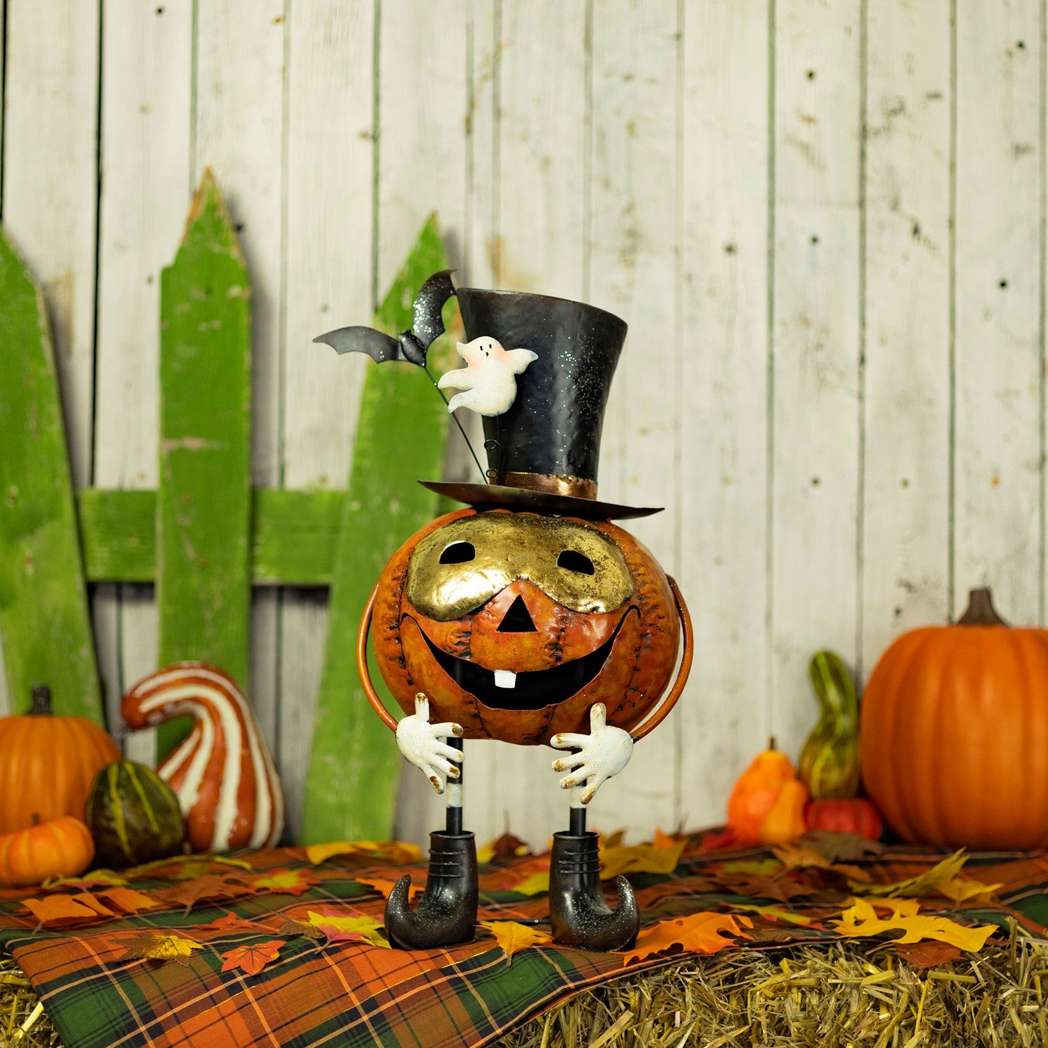 A Halloween decorative pumpkin figure with a gold face, black top hat, white ghost, and black umbrella-shaped legs, posed on a plaid blanket with hay, fall leaves, pumpkins, and a rustic wood plank backdrop.