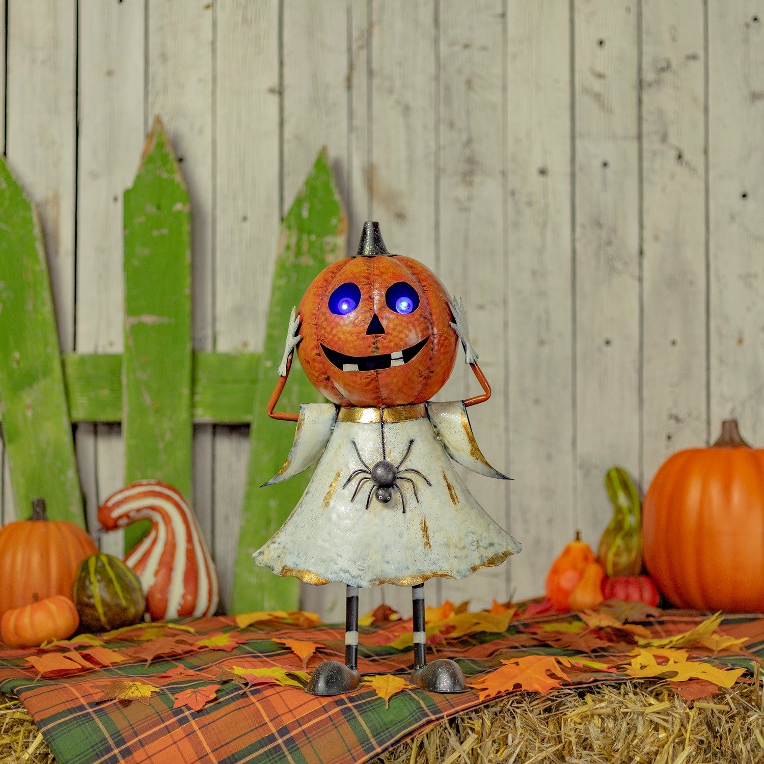 A Halloween metal decoration shaped like a pumpkin-headed figure wearing a distressed white dress, standing on hay with autumn leaves, pumpkins, and a rustic wood plank backdrop.