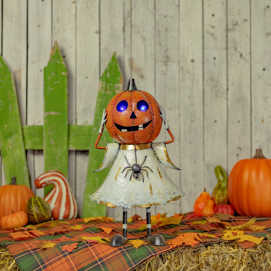 A Halloween metal decoration shaped like a pumpkin-headed figure wearing a distressed white dress, standing on hay with autumn leaves, pumpkins, and a rustic wood plank backdrop.