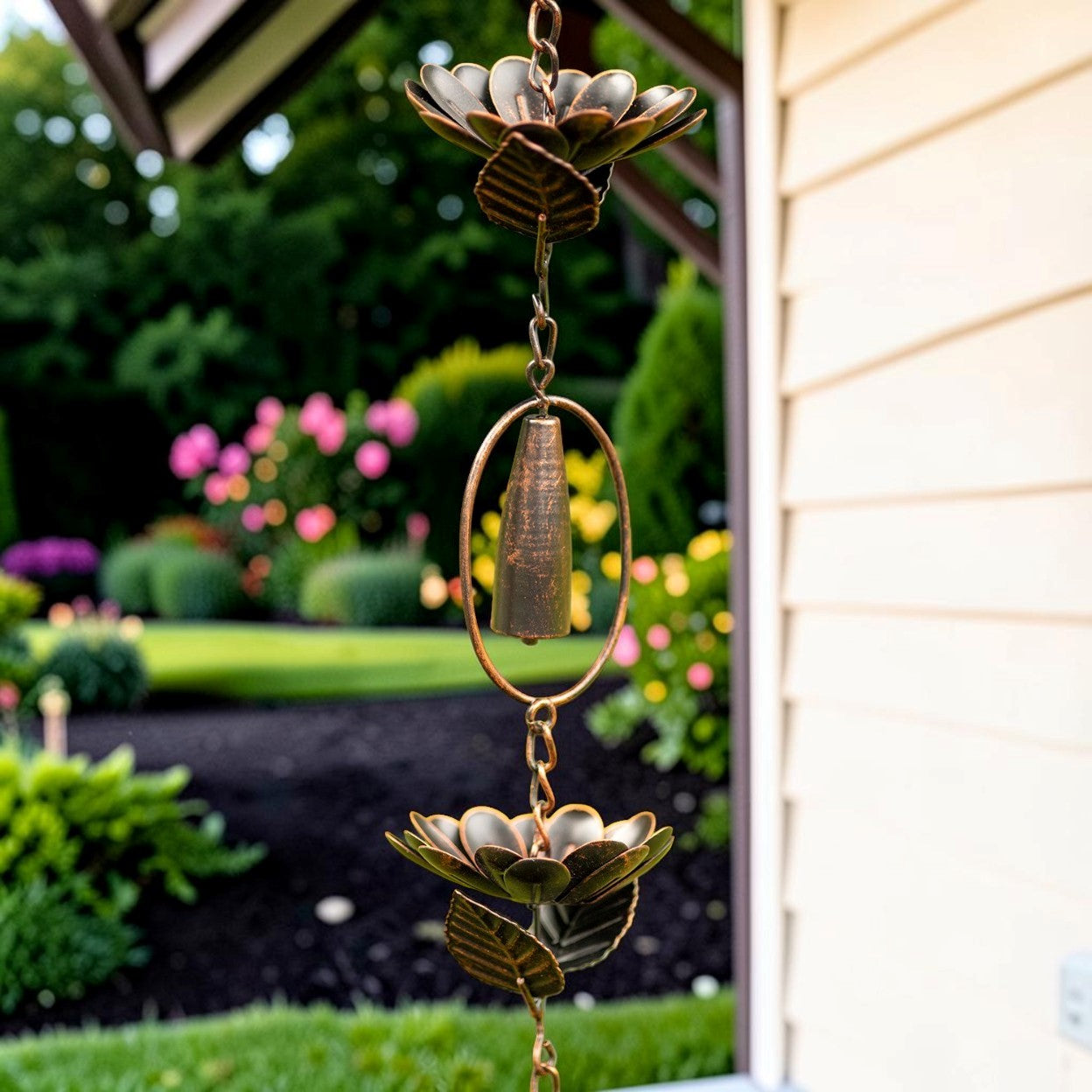 A bronze metal rain garden wind spinner with lotus flower designs hangs from a downspout in a residential garden setting with a blurred background of greenery and flowers.