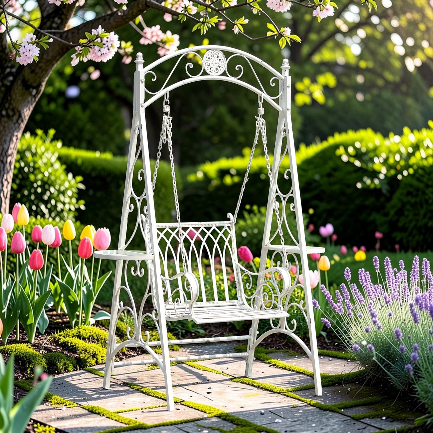 A white wrought-iron garden swing bench placed on a stone path in a lush garden with blooming pink flowers, tulips, lavender, and green hedges in the background.