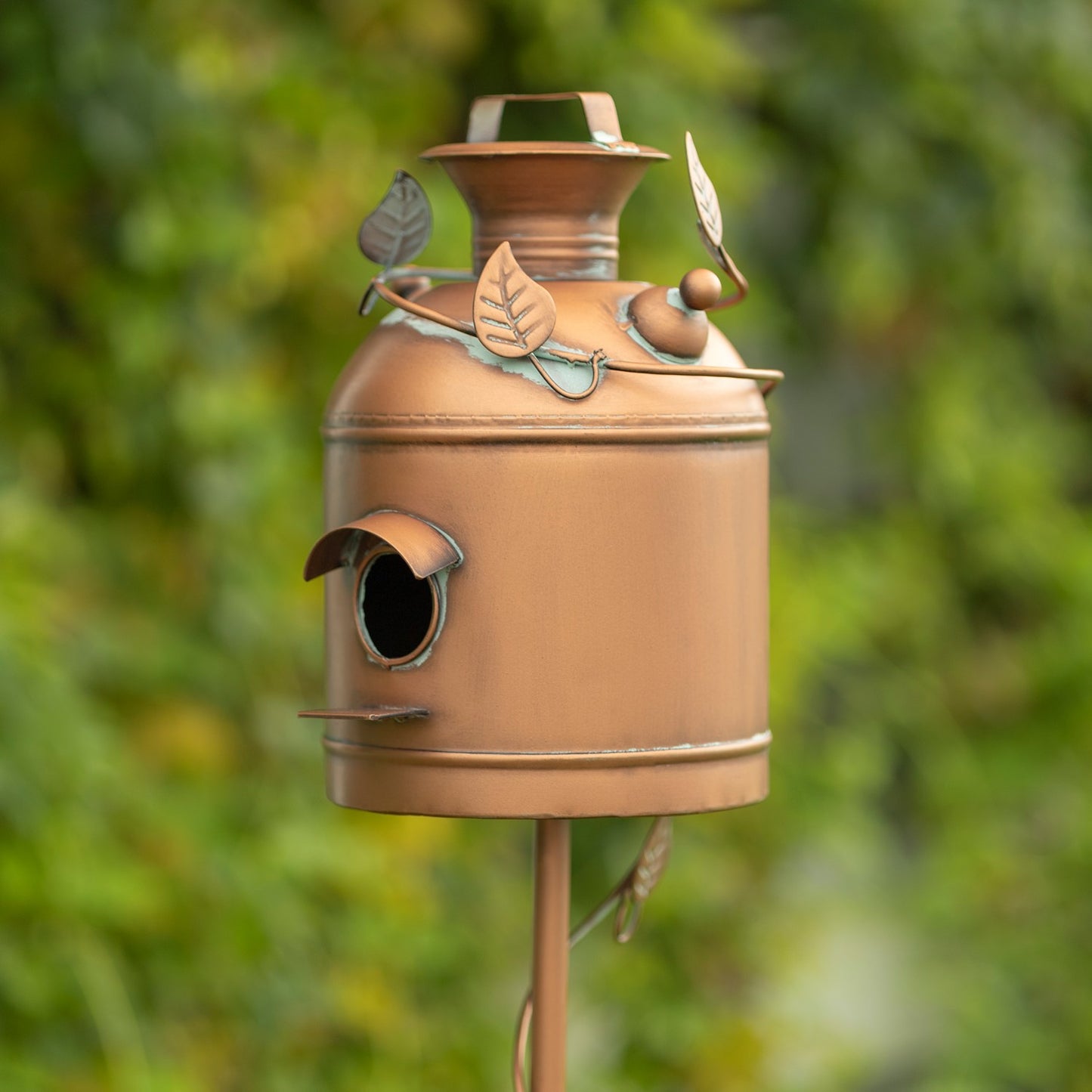 A decorative copper birdhouse with leaf and acorn details, mounted on a metal post against a blurred green garden background.