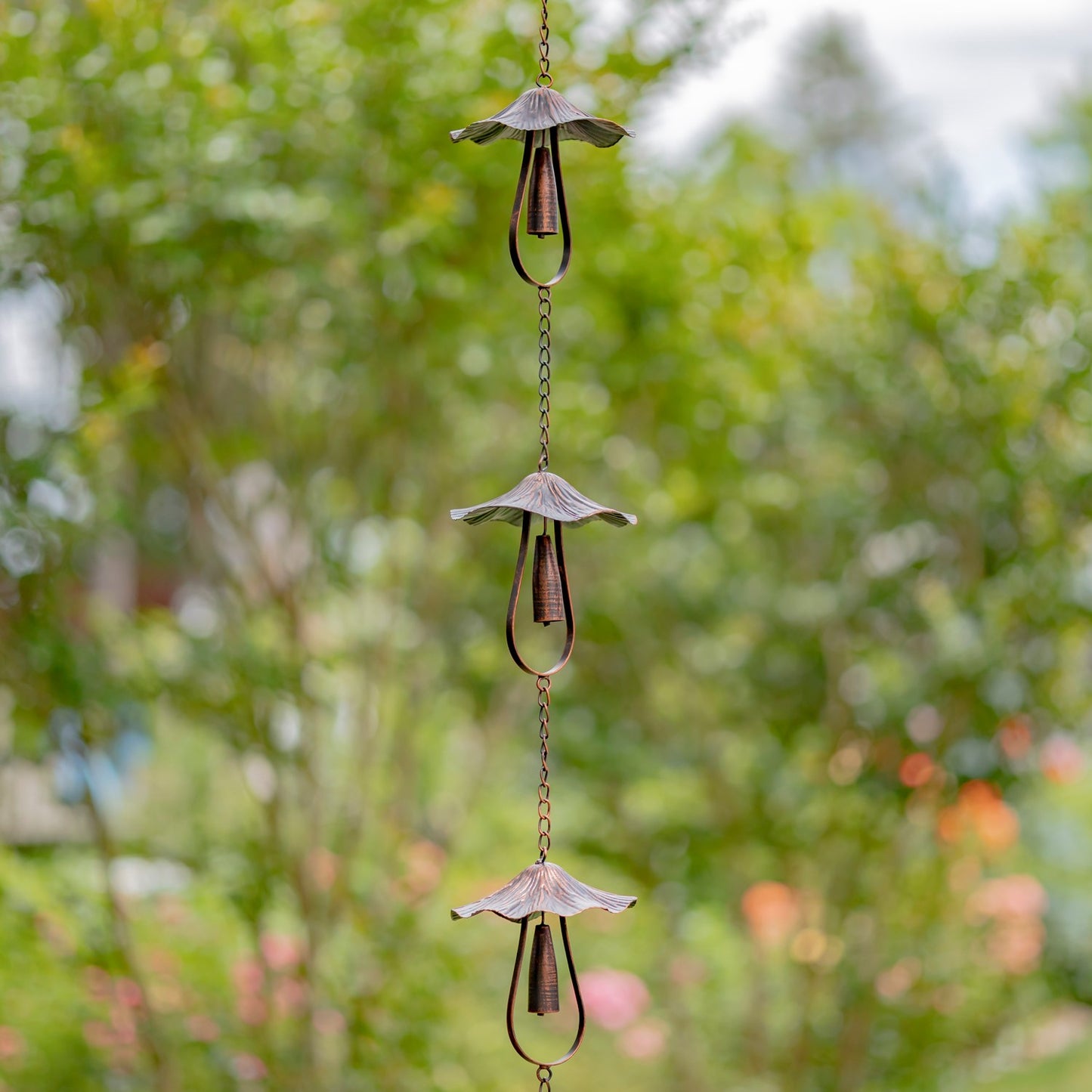 A three-tier metal rain garden chain with mushroom-shaped ornaments hanging outdoors against a blurred green garden background.