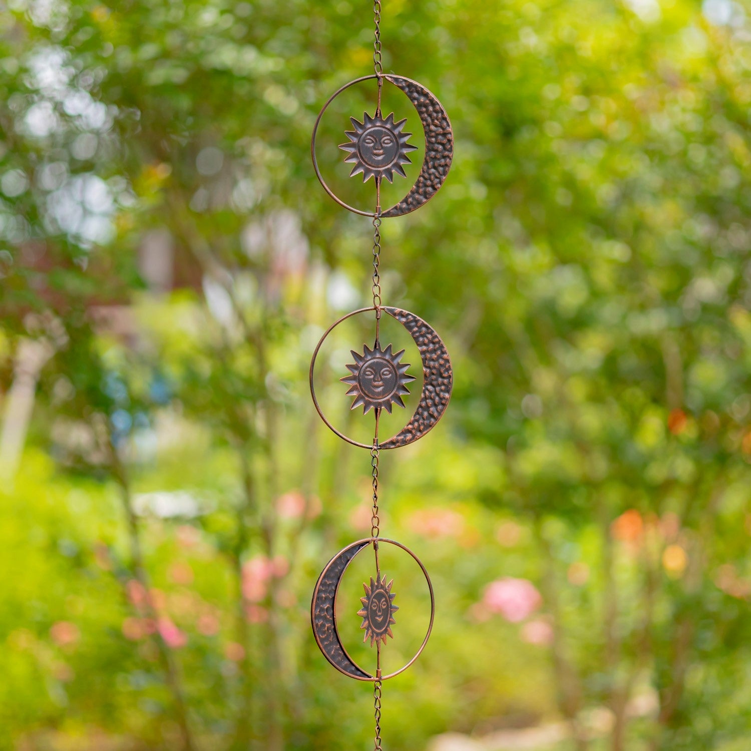 Decorative metal wind chime with sun and moon designs hanging against a blurred green garden background.