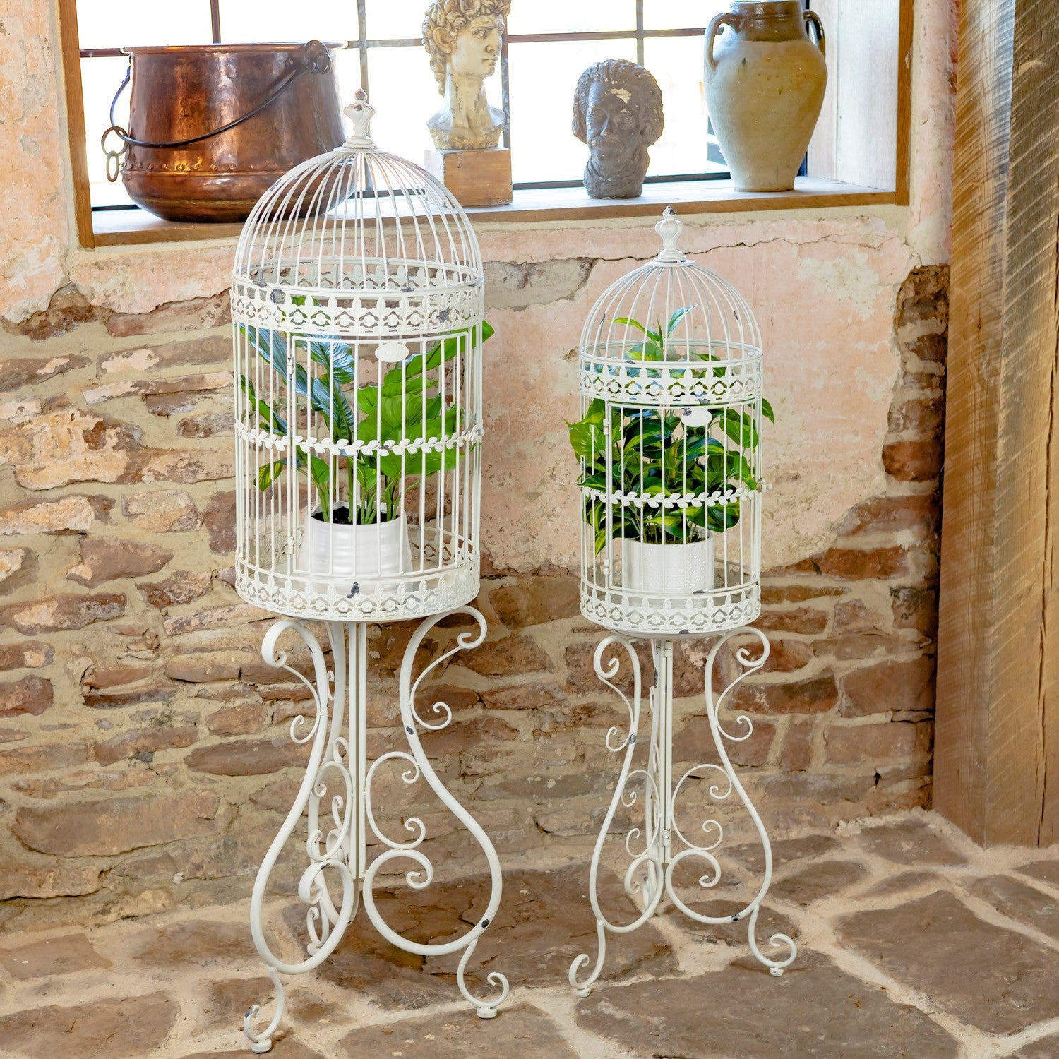 Two white metal birdcage-style plant stands with ornate scroll legs, each holding a green potted plant, displayed against a rustic stone wall and brick floor.