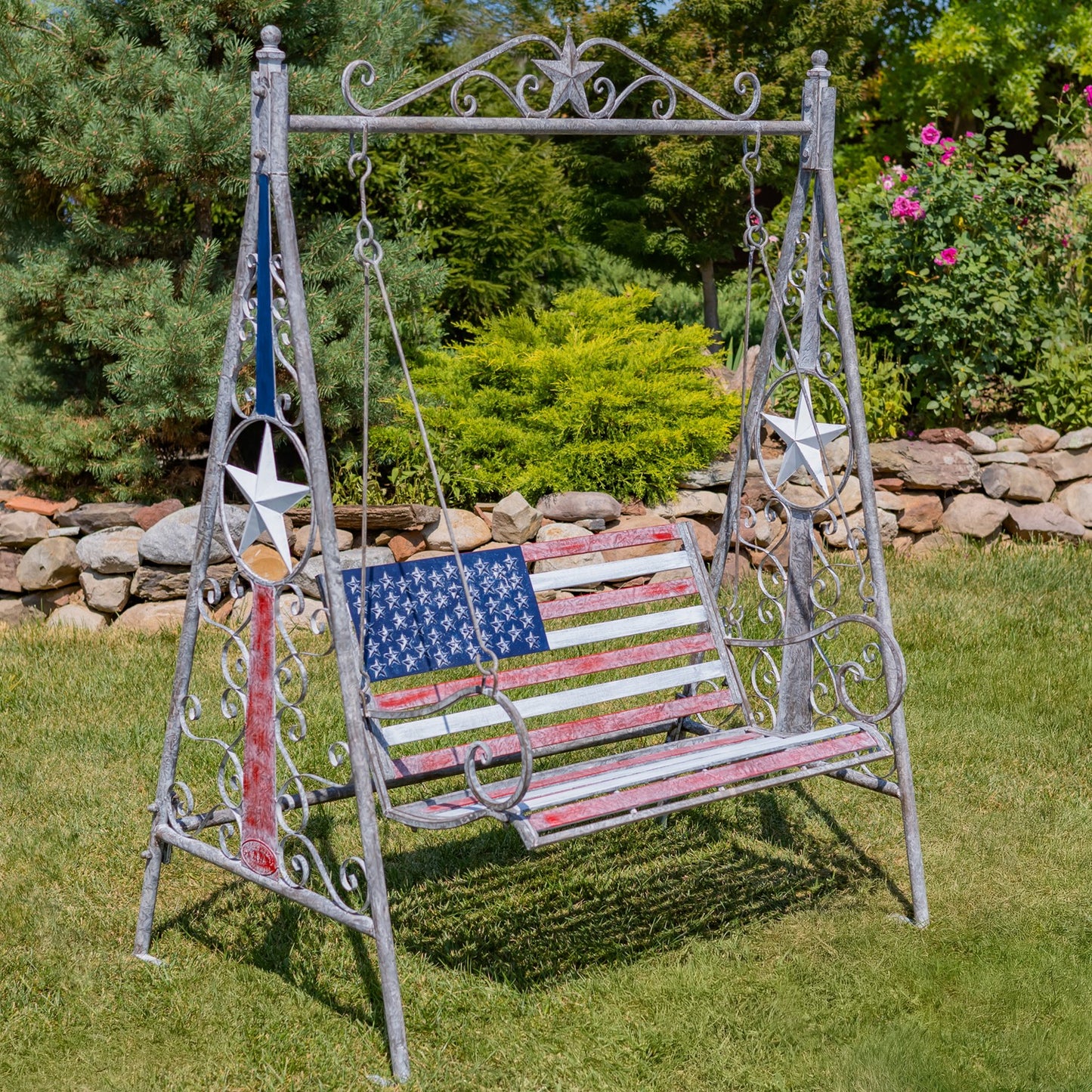 A rustic metal swing bench with an American flag design, set on a green lawn in a garden. The swing has a distressed silver metal frame with a red, white, and blue flag pattern on the seat, and a vertical Texas flag hanging on the left side.