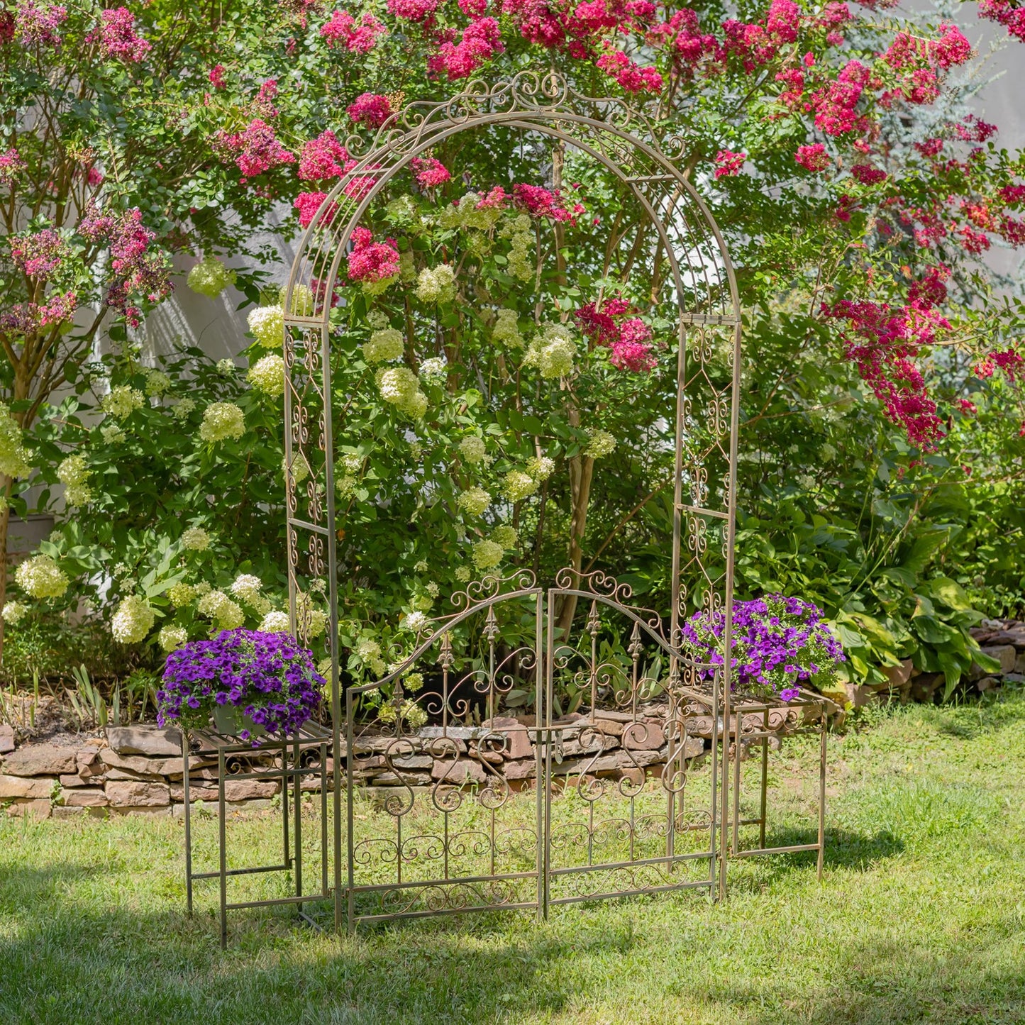 An ornate metal garden bench with an arched trellis design, set in a lush garden with pink and white hydrangeas and purple flowers.
