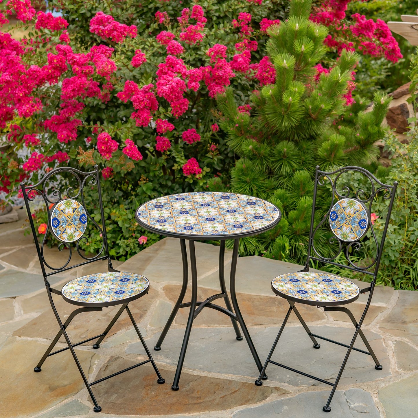 A three-piece outdoor bistro set featuring a round table and two matching chairs with black metal frames and colorful mosaic tile seats and backs, set on a stone patio in front of a lush garden of pink flowers and green foliage.
