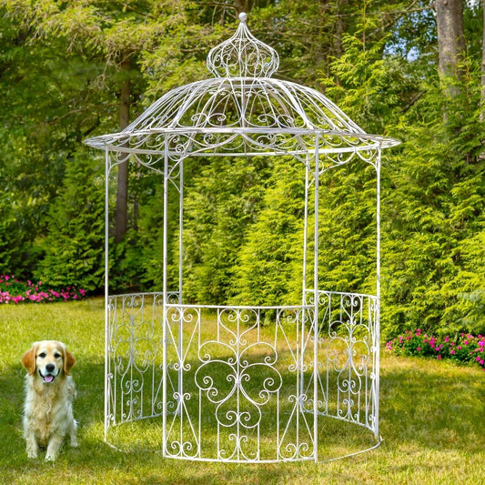 A white wrought-iron garden gazebo with an ornate pointed roof and decorative scrollwork, shown outdoors on a grassy lawn with a small dog standing nearby and a background of green trees and shrubs.