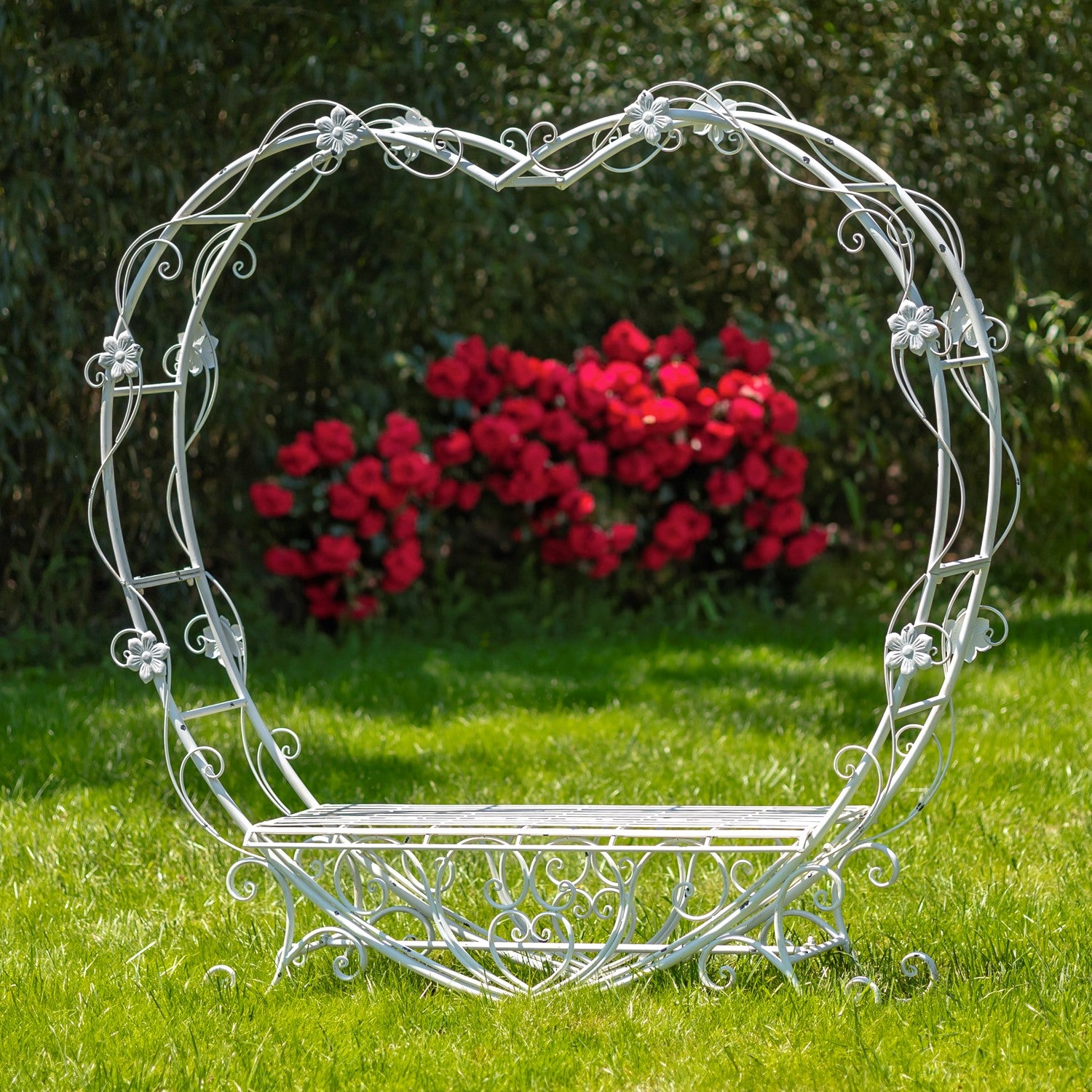 Decorative white metal heart-shaped archway in a garden setting with red flowers in the background.
