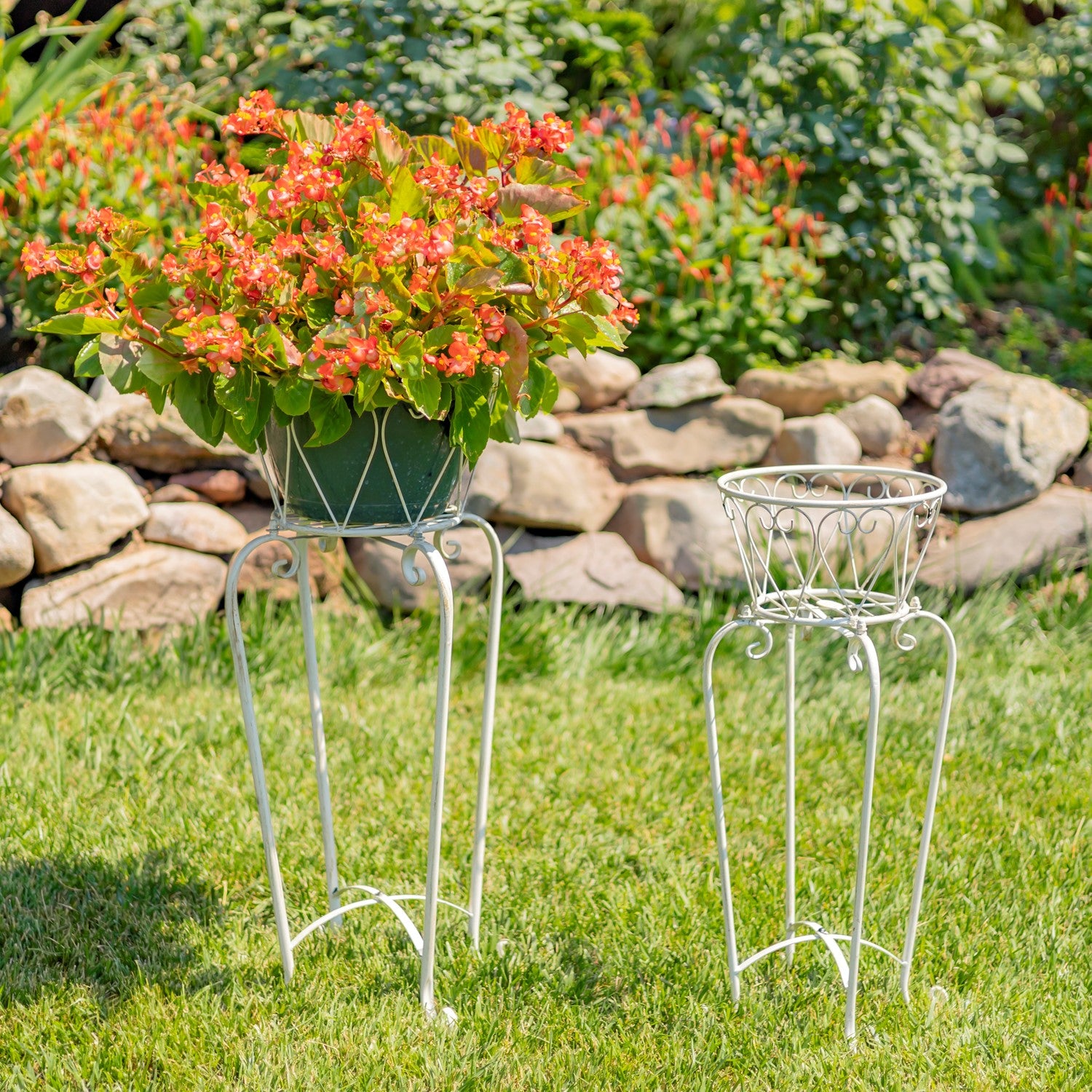Two white metal garden plant stands on a grassy lawn, with a potted flowering plant on the taller stand and an empty planter basket on the shorter stand. A stone garden bed and green foliage are in the background.