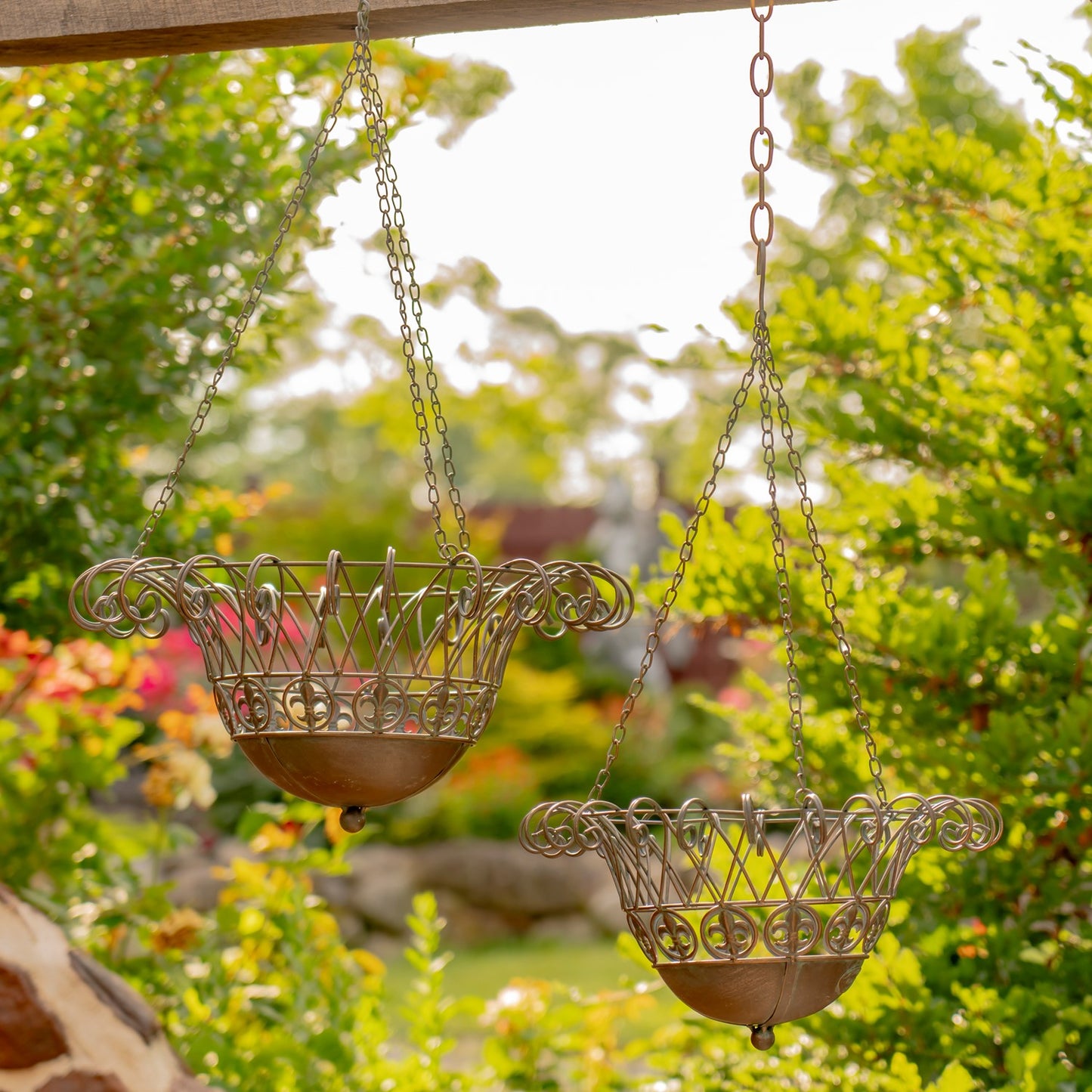 Two ornate metal hanging planters with scrollwork details, suspended by chains from a wooden beam in a sunlit garden setting.