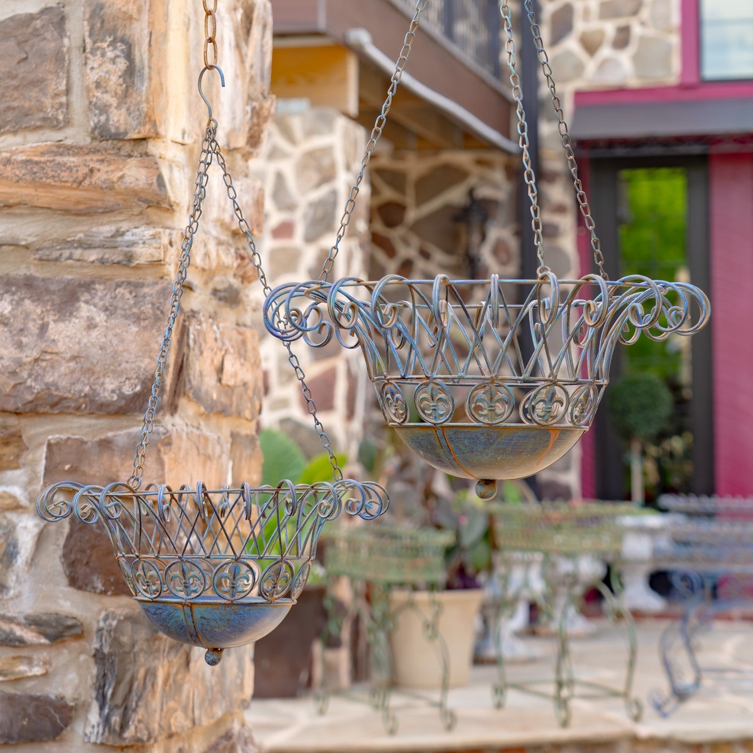 Two ornate hanging metal planters with scrollwork details and blue-gray patina, suspended by chains outdoors against a stone wall and patio background.