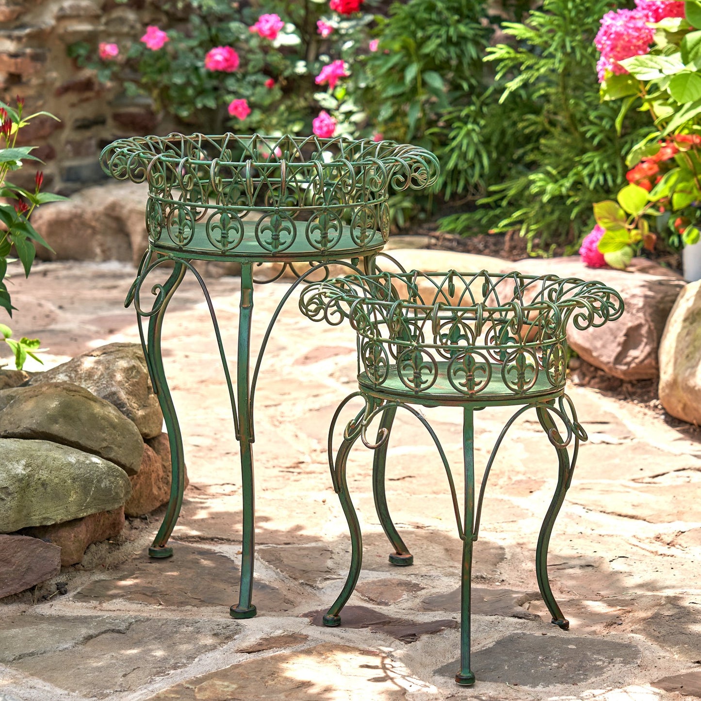 Two green metal garden plant stands with ornate scrollwork, one tall and one short, placed on a stone patio in a garden with flowers and rocks in the background.