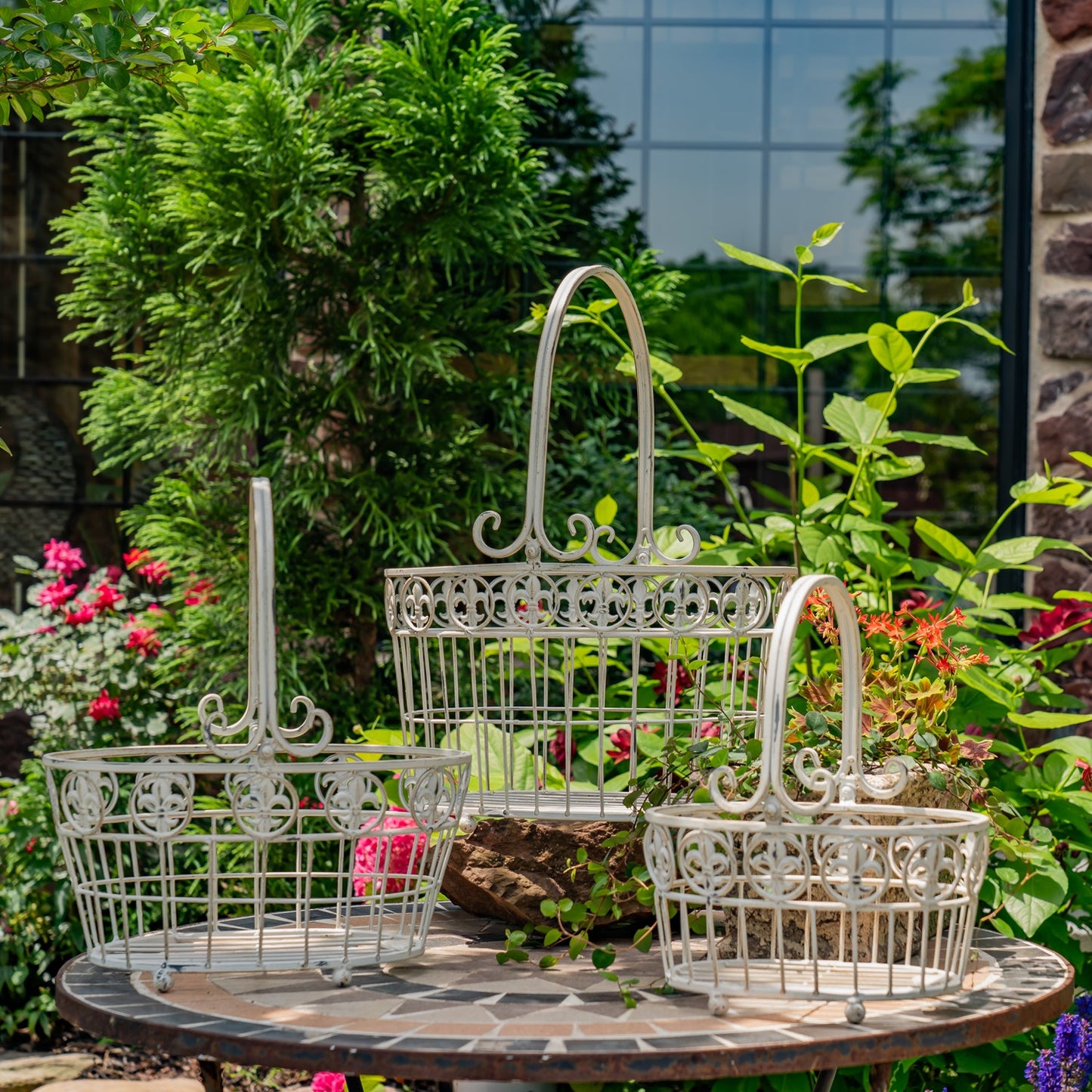 Three white ornate metal garden baskets of different sizes arranged on a round stone table in a garden setting with green plants and flowers.