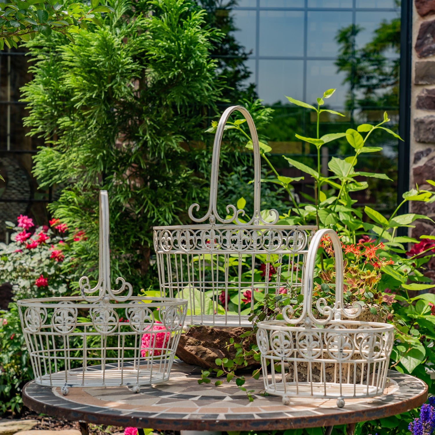 Three white ornate metal garden baskets of different sizes arranged on a round stone table in a garden setting with green plants and flowers.
