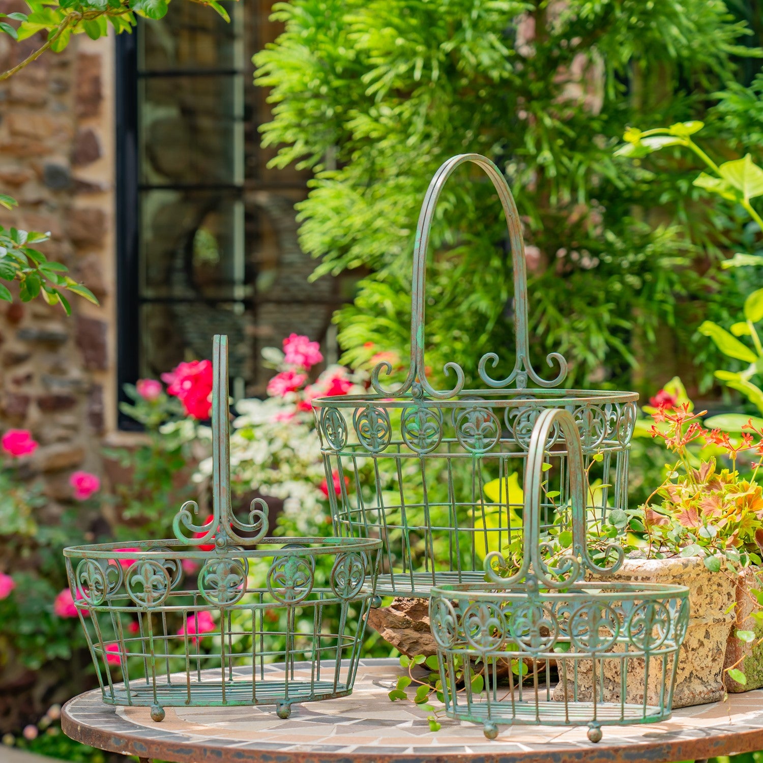 Three green metal wire baskets with decorative scrollwork and arched handles, displayed on a stone table in a garden setting with flowers and foliage.