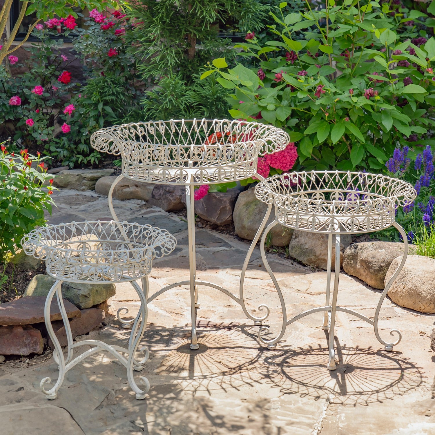 Three white metal garden plant stands arranged on a stone patio in a lush garden setting with green foliage and pink flowers.