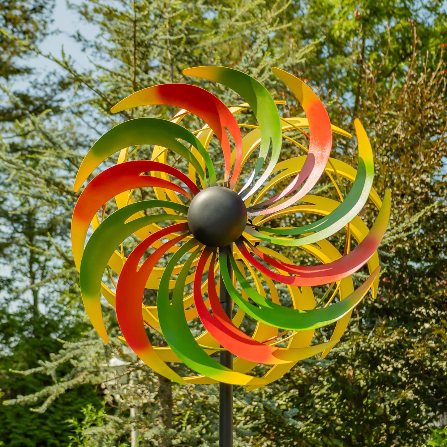 A colorful metal wind spinner with alternating red, yellow, and green curved blades and a dark gray central hub, mounted on a black pole outdoors against a background of green trees.