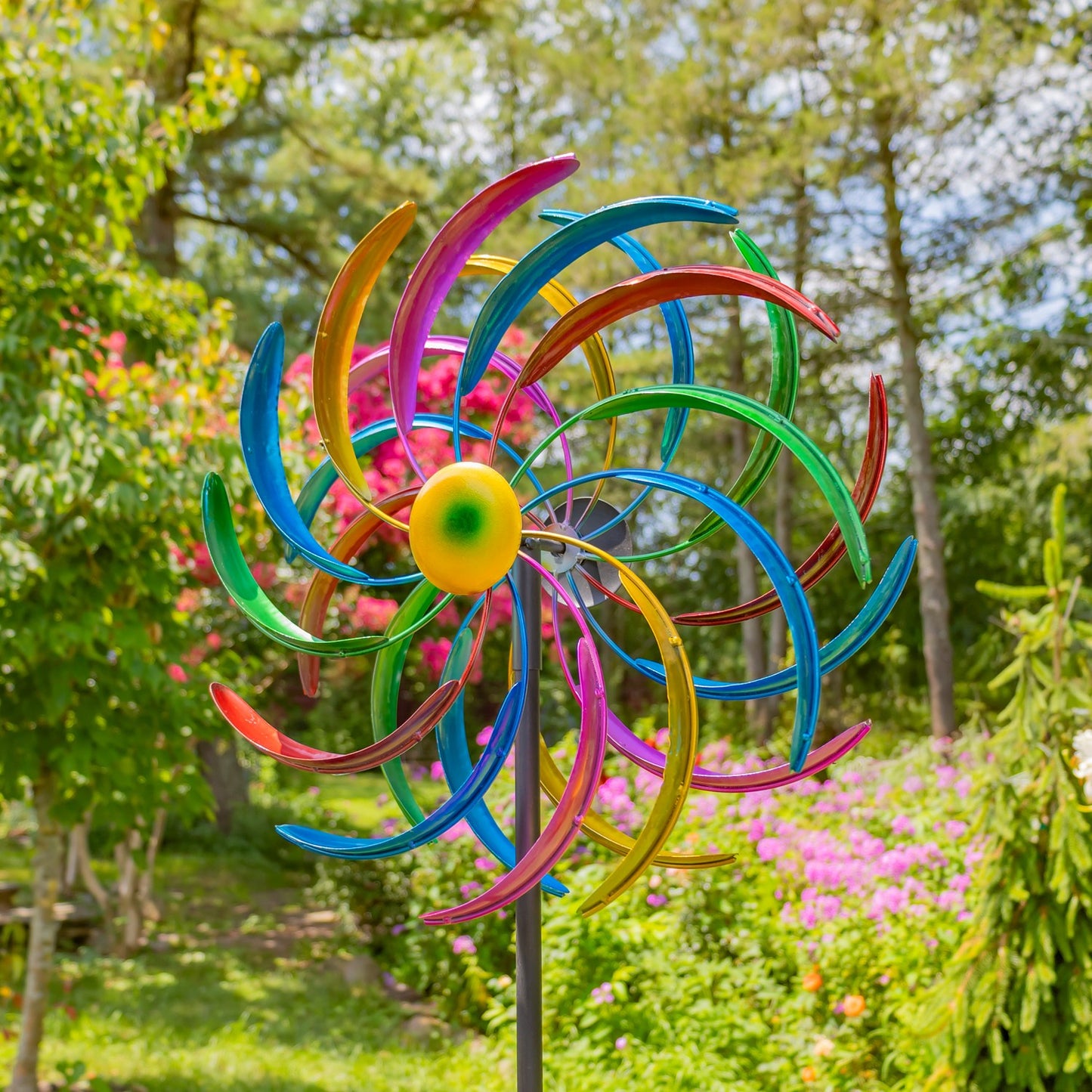A colorful metal garden wind spinner with multiple curved blades in blue, green, pink, yellow, and orange, mounted on a metal stand in a garden setting with trees and flowers in the background.