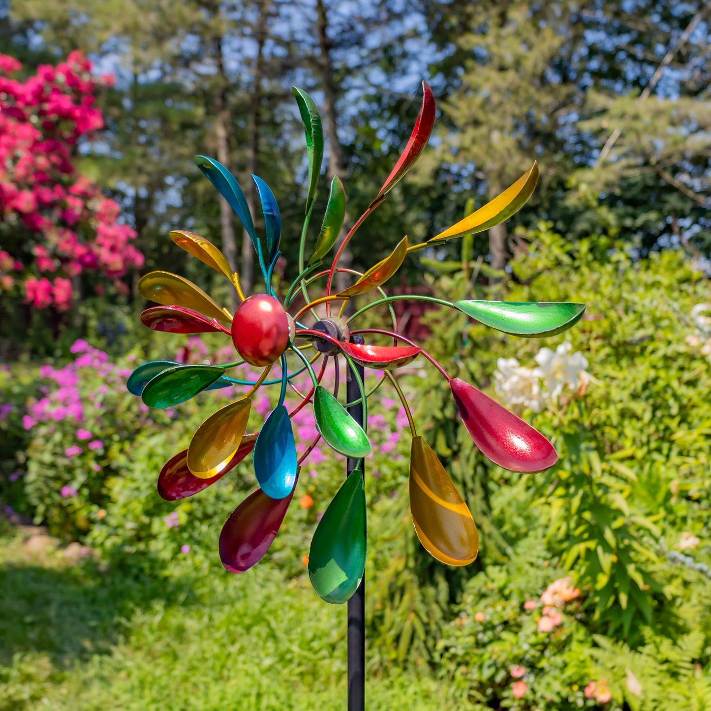 A colorful metal garden wind spinner with vibrant, rainbow-colored petal-like blades and a red center, mounted on a black stake in a lush green garden.