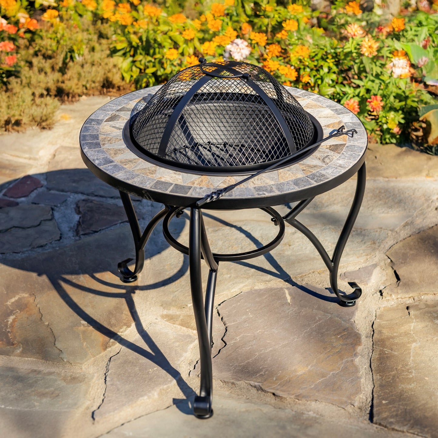 Outdoor round fire pit table with a mosaic tile rim, black metal legs, and a black mesh fire dome cover, set on a stone patio with flowers in the background.