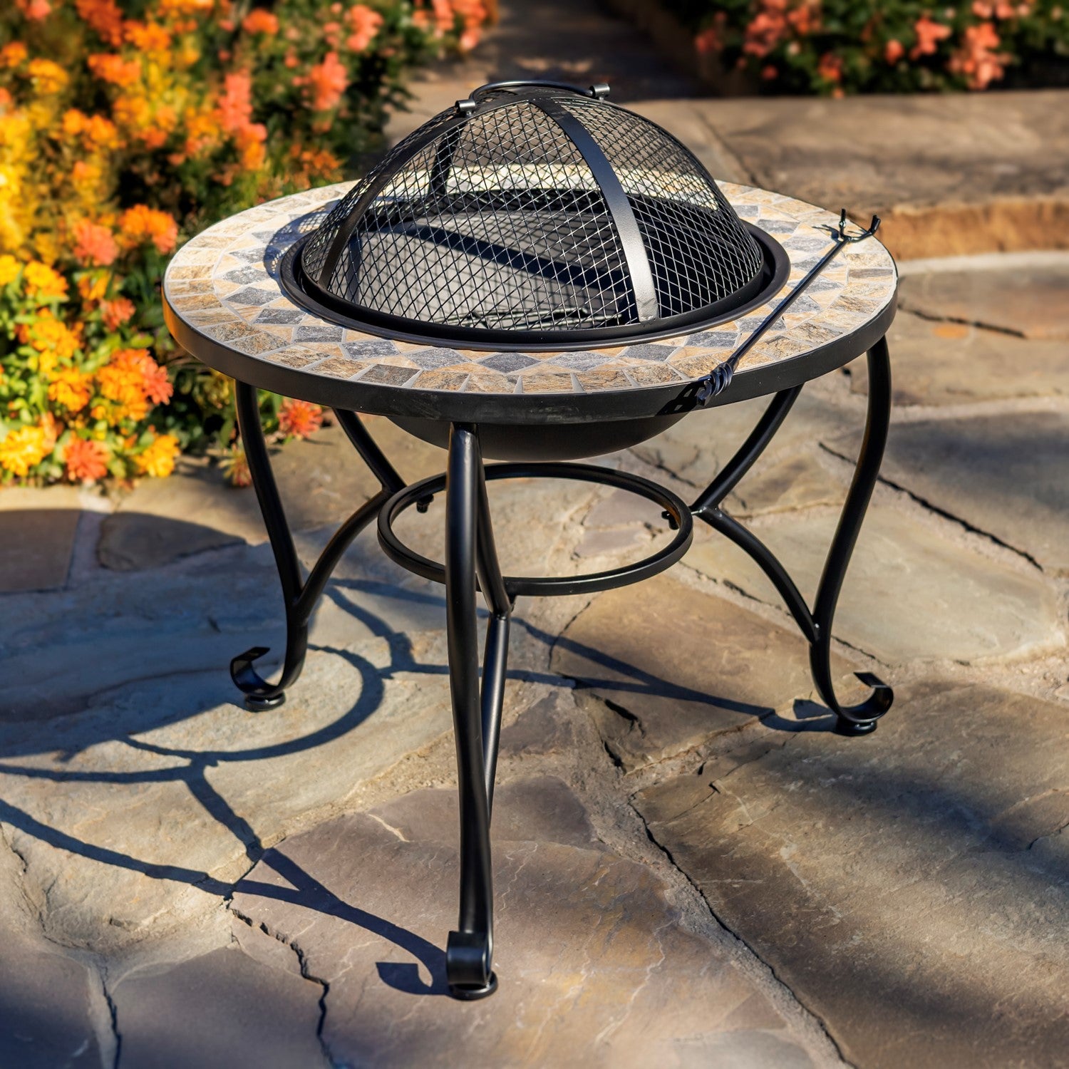 Outdoor round fire pit table with a mosaic stone-look top and black metal legs, shown on a stone patio with flowers in the background.