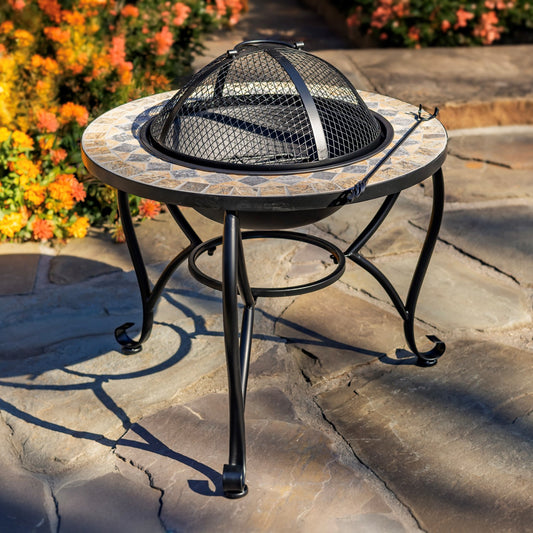 Outdoor round fire pit table with a mosaic stone-look top and black metal legs, shown on a stone patio with flowers in the background.