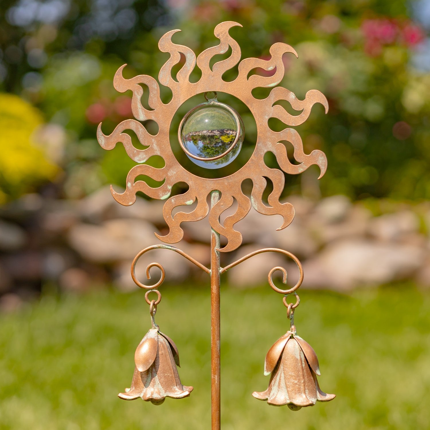 A decorative metal garden stake shaped like a sun with two hanging bell-shaped ornaments, photographed outdoors on a blurred green lawn and garden background.