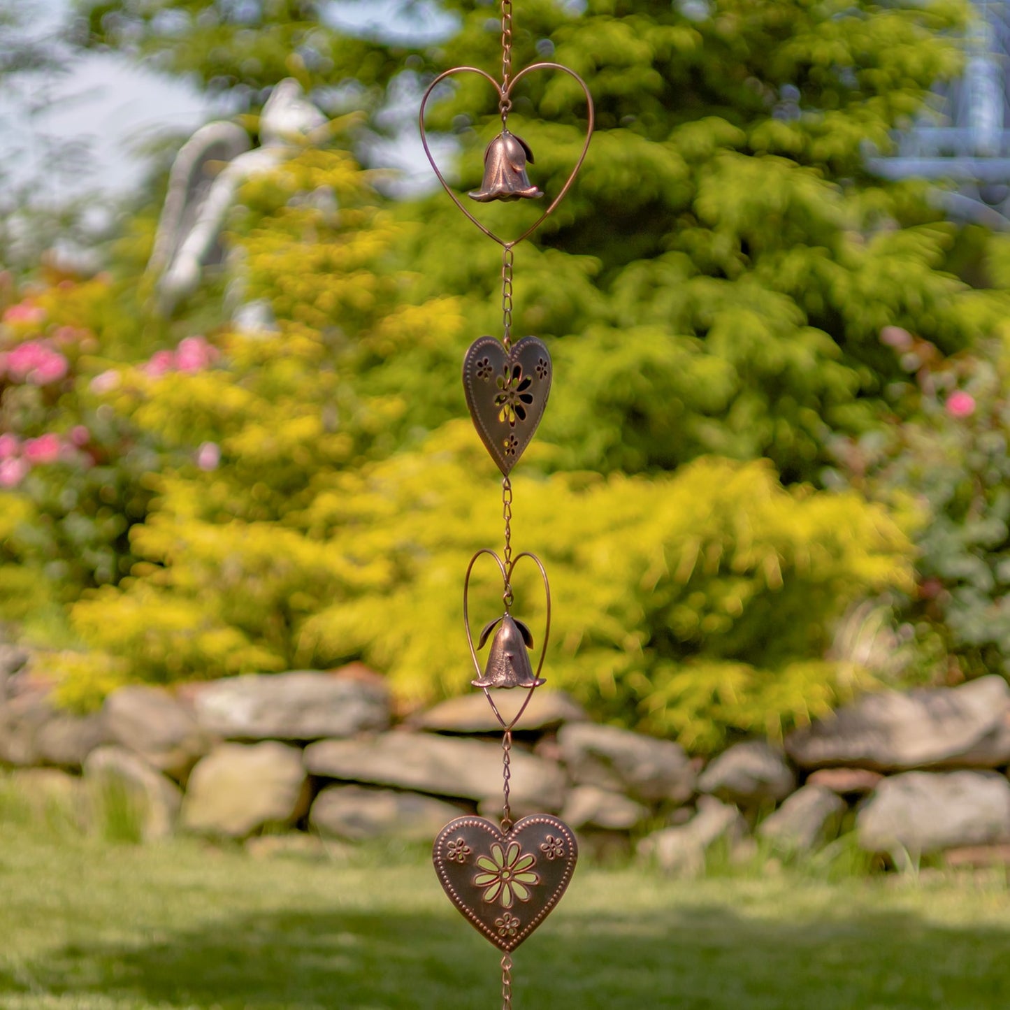 A copper-toned metal garden wind spinner featuring a bell-shaped flower suspended inside a heart-shaped wire frame, set against a blurred green grass background.