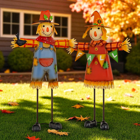 Two scarecrows with autumn-themed hats and scarves standing on grass with a house and trees in the background.
