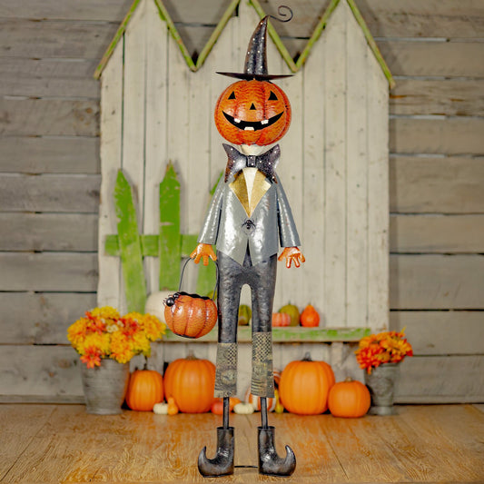A Halloween decorative scarecrow figure with a carved orange jack-o'-lantern head, black witch hat, silver-gray suit, and orange pumpkin basket, standing in front of a rustic wood backdrop with pumpkins and flowers.