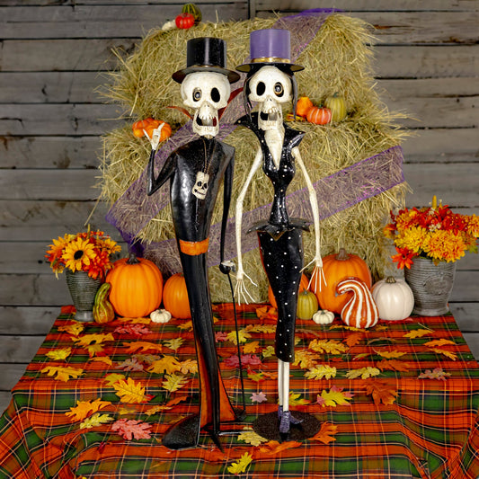 Two decorative skeleton figurines dressed in black formal attire stand on a plaid autumn tablecloth with pumpkins, fall leaves, flowers, and a hay bale backdrop.