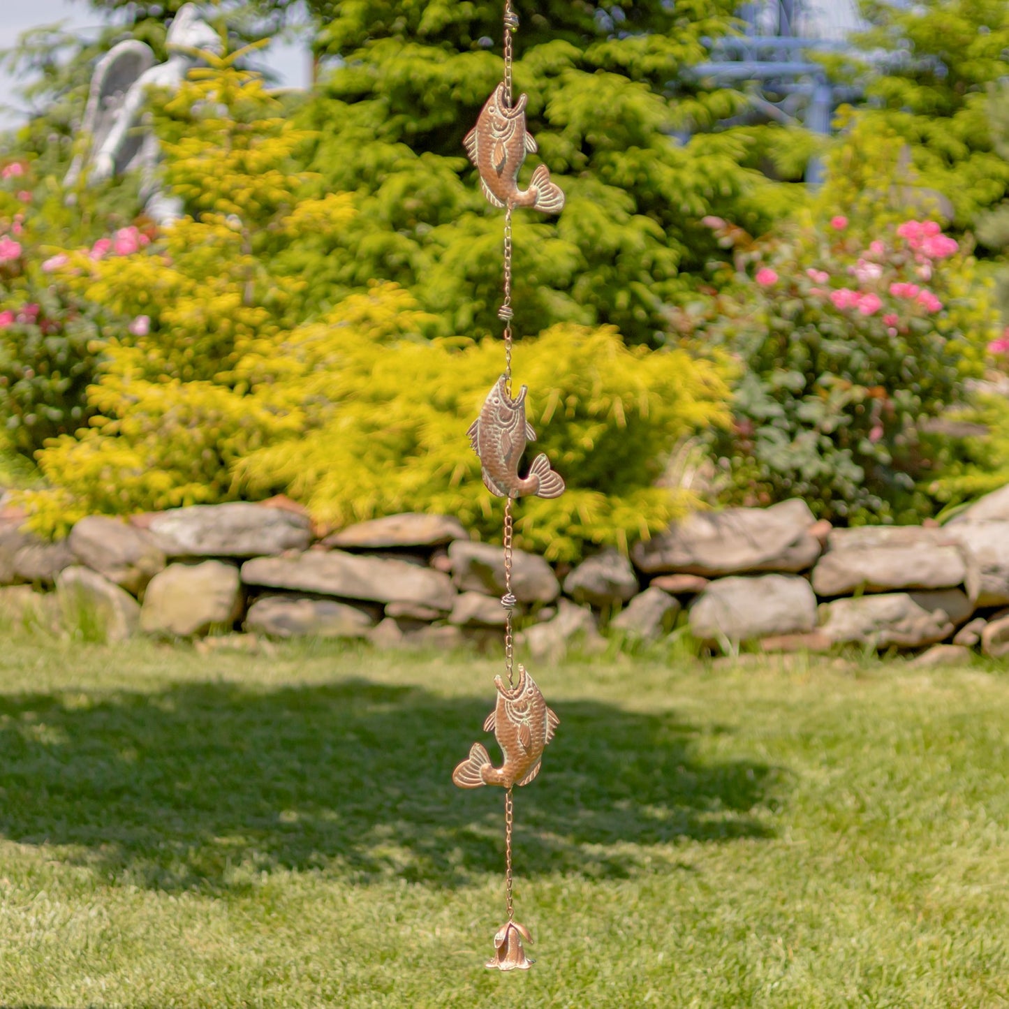 A vertical hanging wind spinner ornament featuring three detailed fish figures on a chain, set against a blurred garden background with green grass, bushes, and flowers.