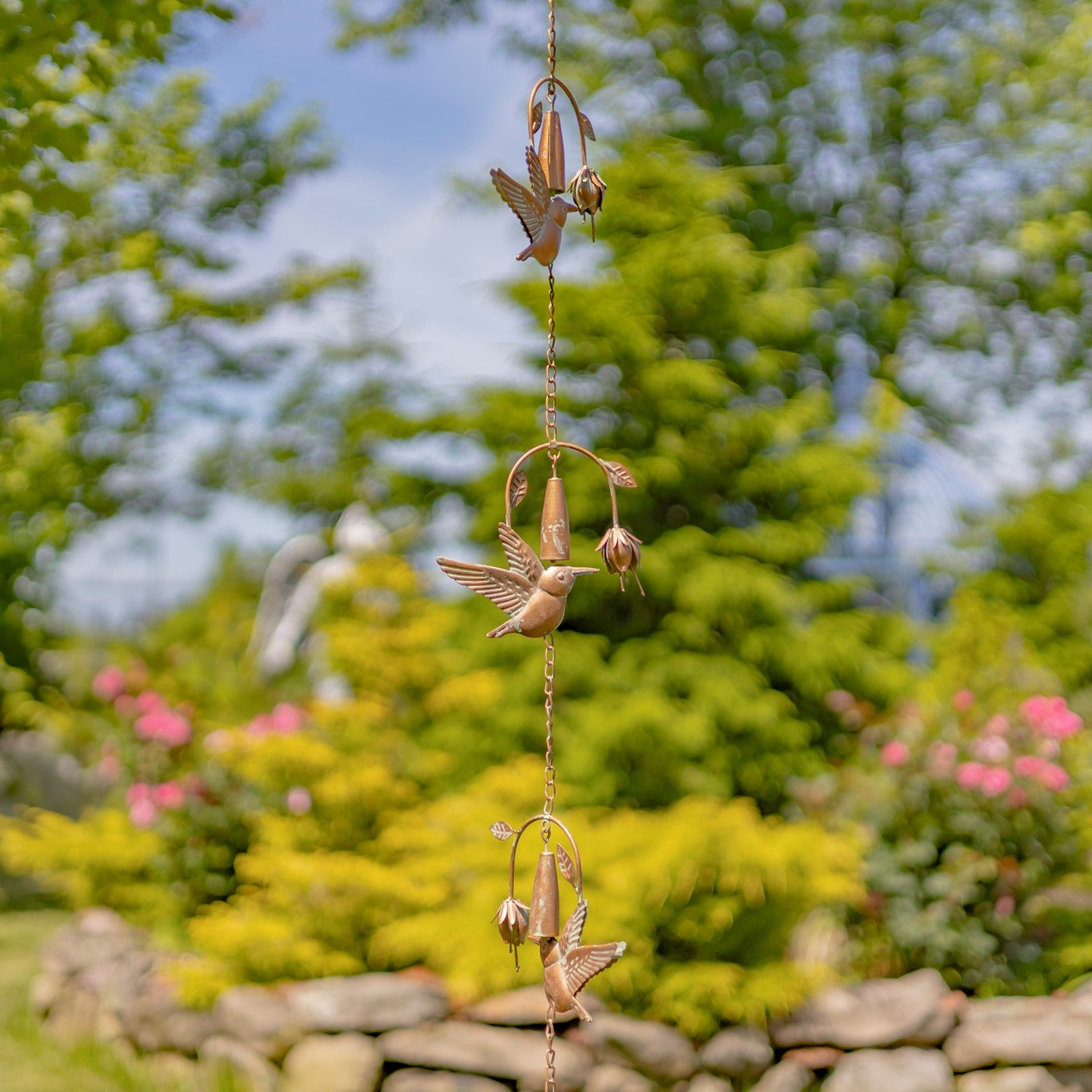A copper hummingbird garden hanging ornament with three detailed copper hummingbirds and flower buds suspended from a wire, set against a blurred green garden background.