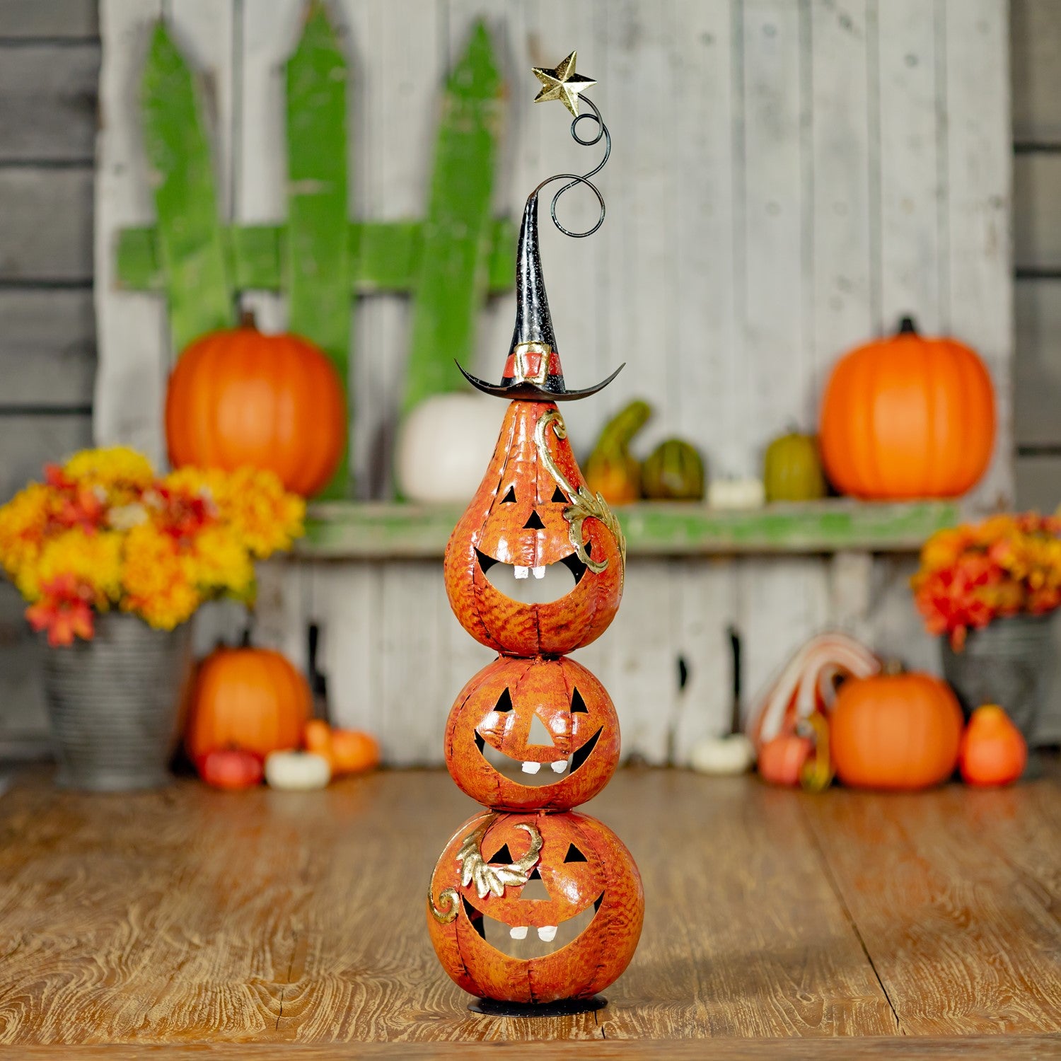 A decorative stacked jack-o'-lantern tower centerpiece in orange glass, topped with a black witch hat and a small gold star, displayed on a wooden surface with pumpkins and autumn flowers in the background.