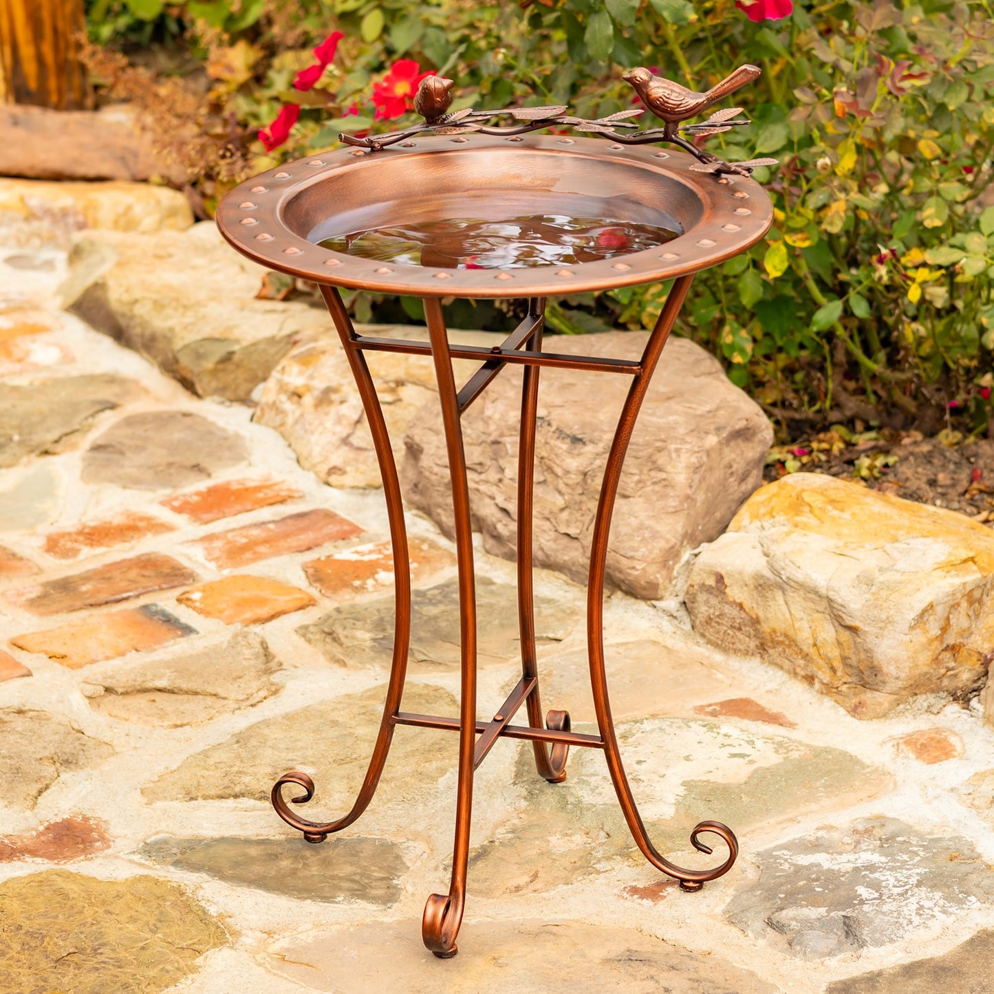 Outdoor copper-finished bird bath with a round tray, decorative bird perches on the rim, and a curved metal stand on a stone patio with garden plants in the background.