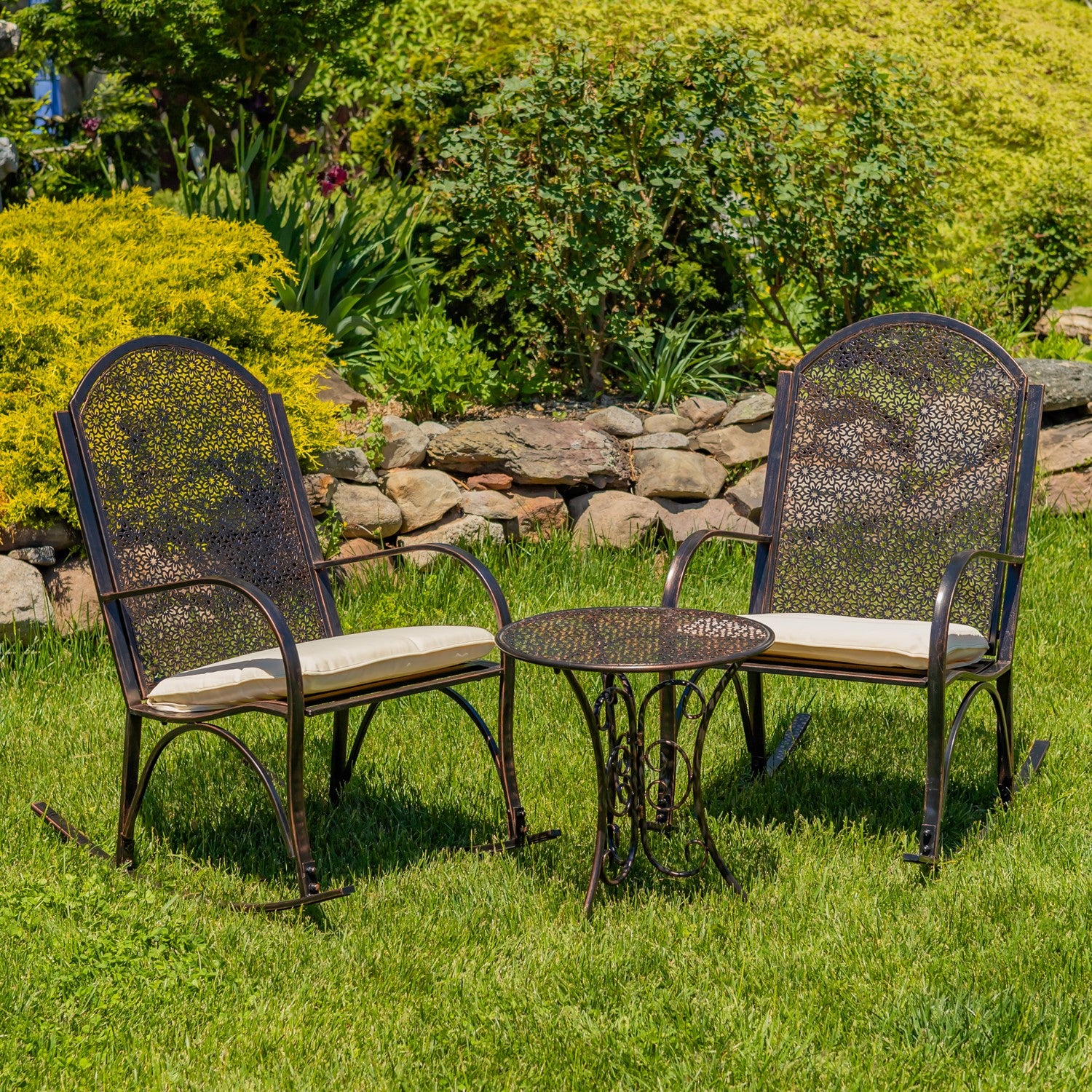 A 3-piece outdoor patio set featuring two dark brown metal chairs with cream cushions and a matching small round table, set on a green lawn in front of a stone wall and garden foliage.