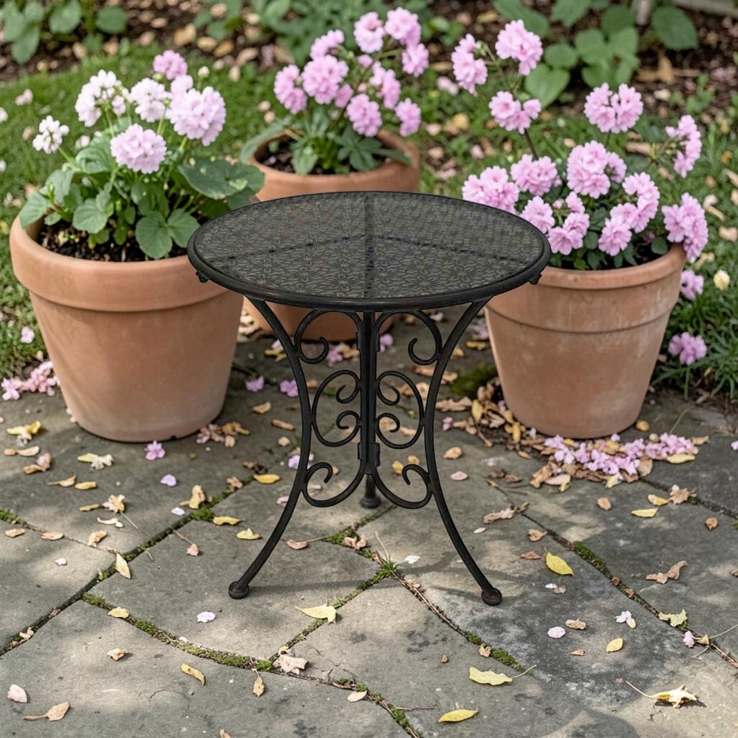 A round black metal patio bistro table with a mesh top and ornate scrollwork legs, set on a stone patio between three potted pink flowers.