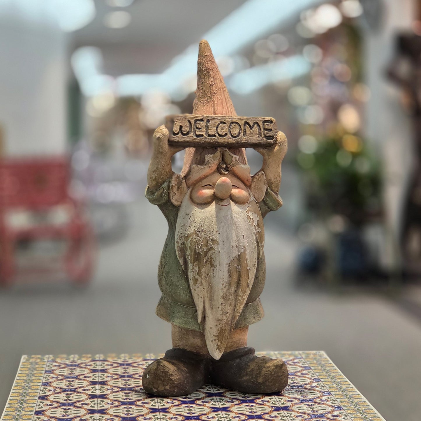 A rustic, weathered gnome garden statue holding a 'WELCOME' sign, displayed on a patterned tile surface with a blurred indoor background.